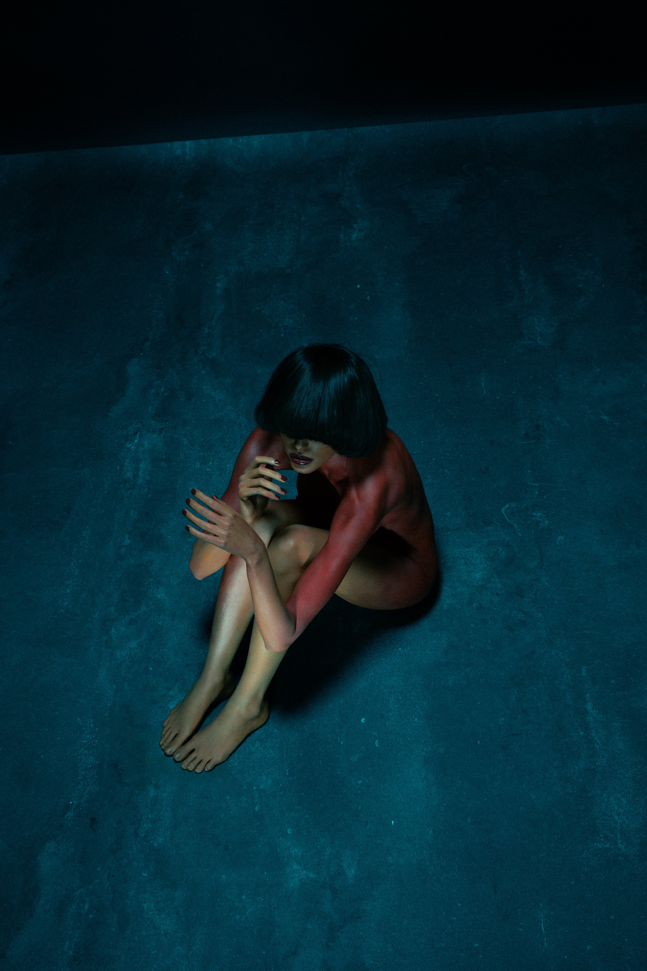 A woman with black hair and light skin, sitting on a dark, textured floor, with her knees pulled close to her chest. She is topless, with her arms crossed over her knees and her head bowed down, obscuring her face.