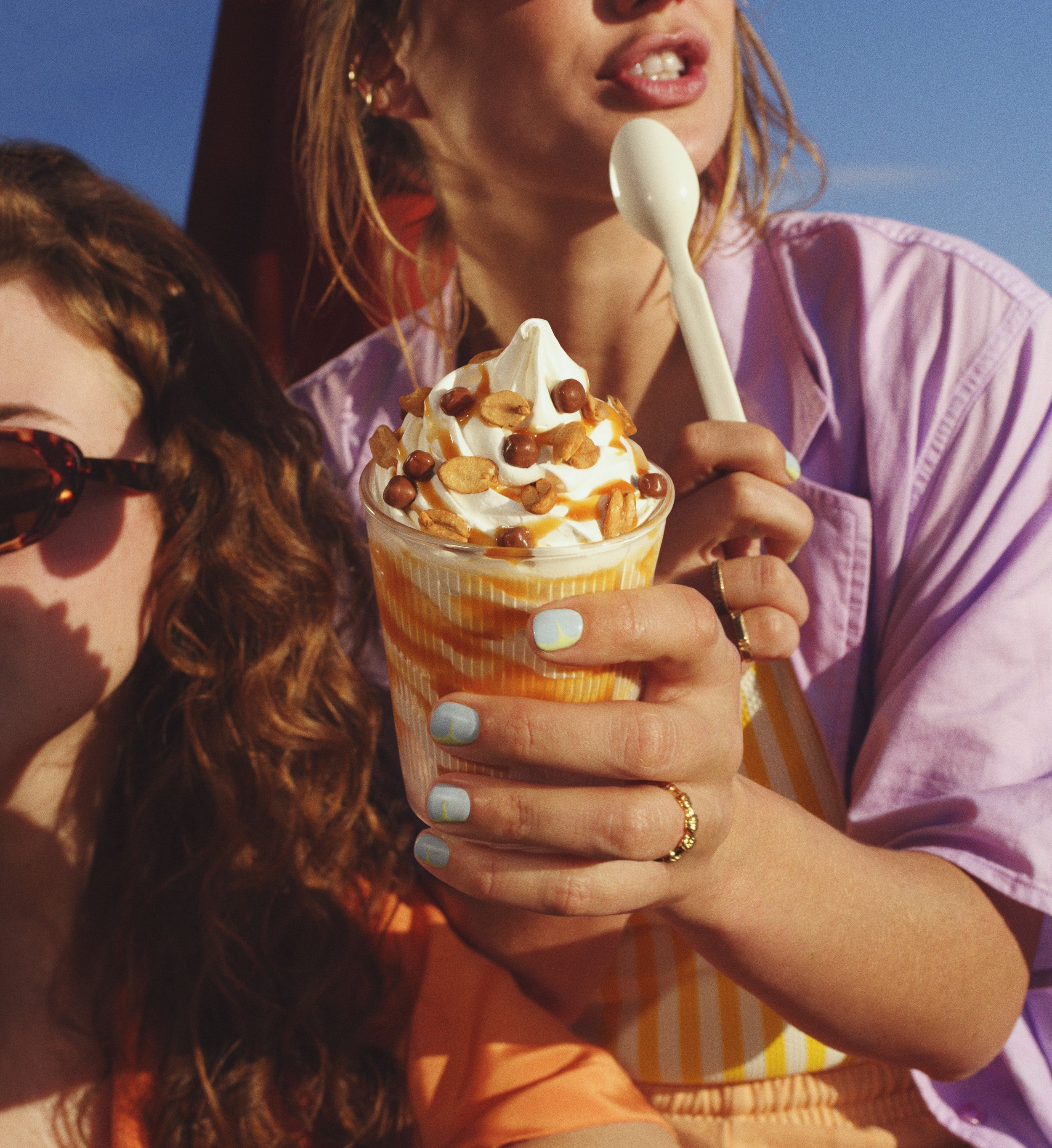 Close-up of a woman holding a cup of soft-serve ice cream topped with caramel syrup, peanuts, and chocolate balls, with two women partially visible beside her, one wearing sunglasses.