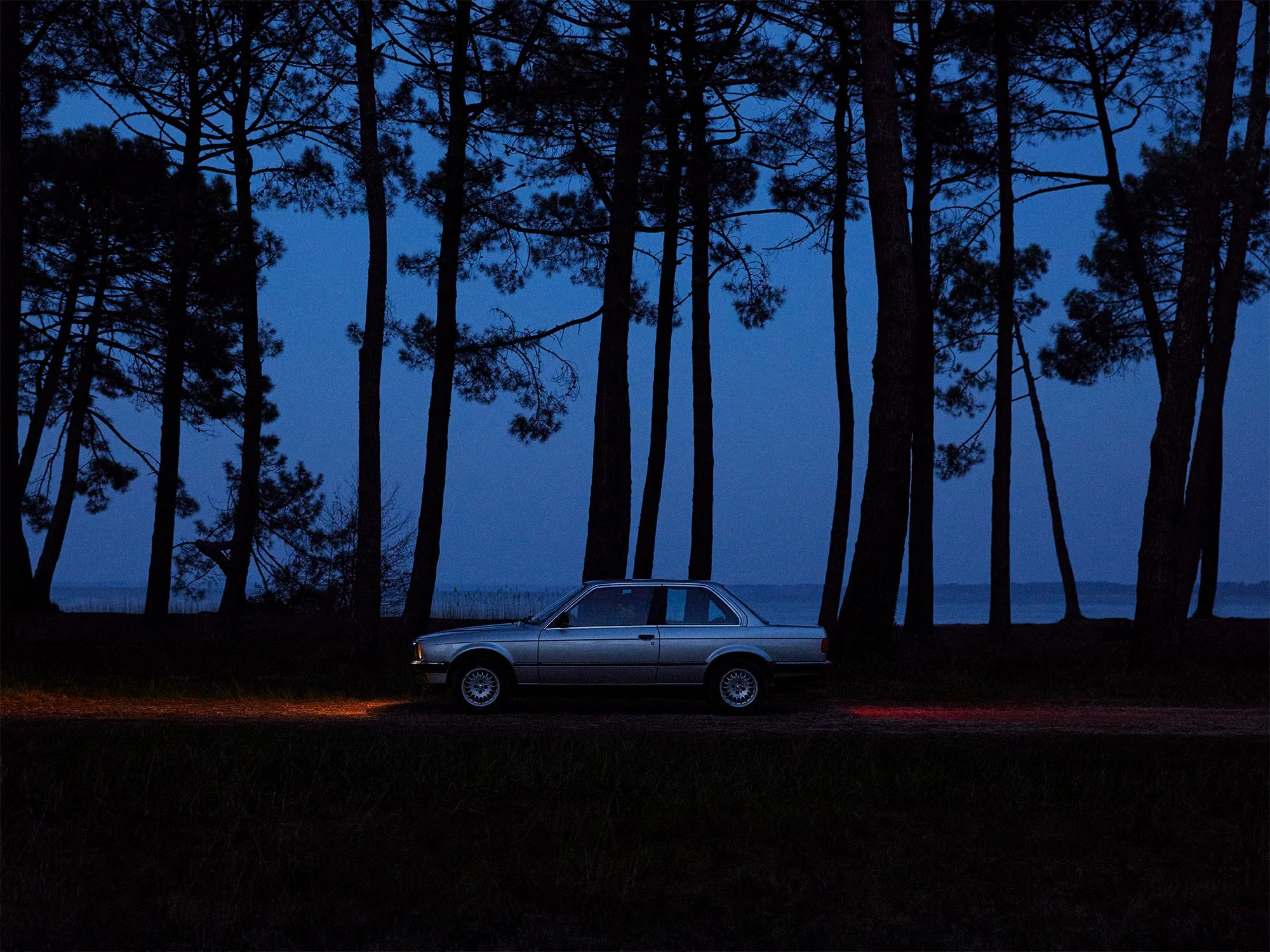 A silver car parked on a dirt road at dusk, surrounded by tall trees with a body of water visible in the background.