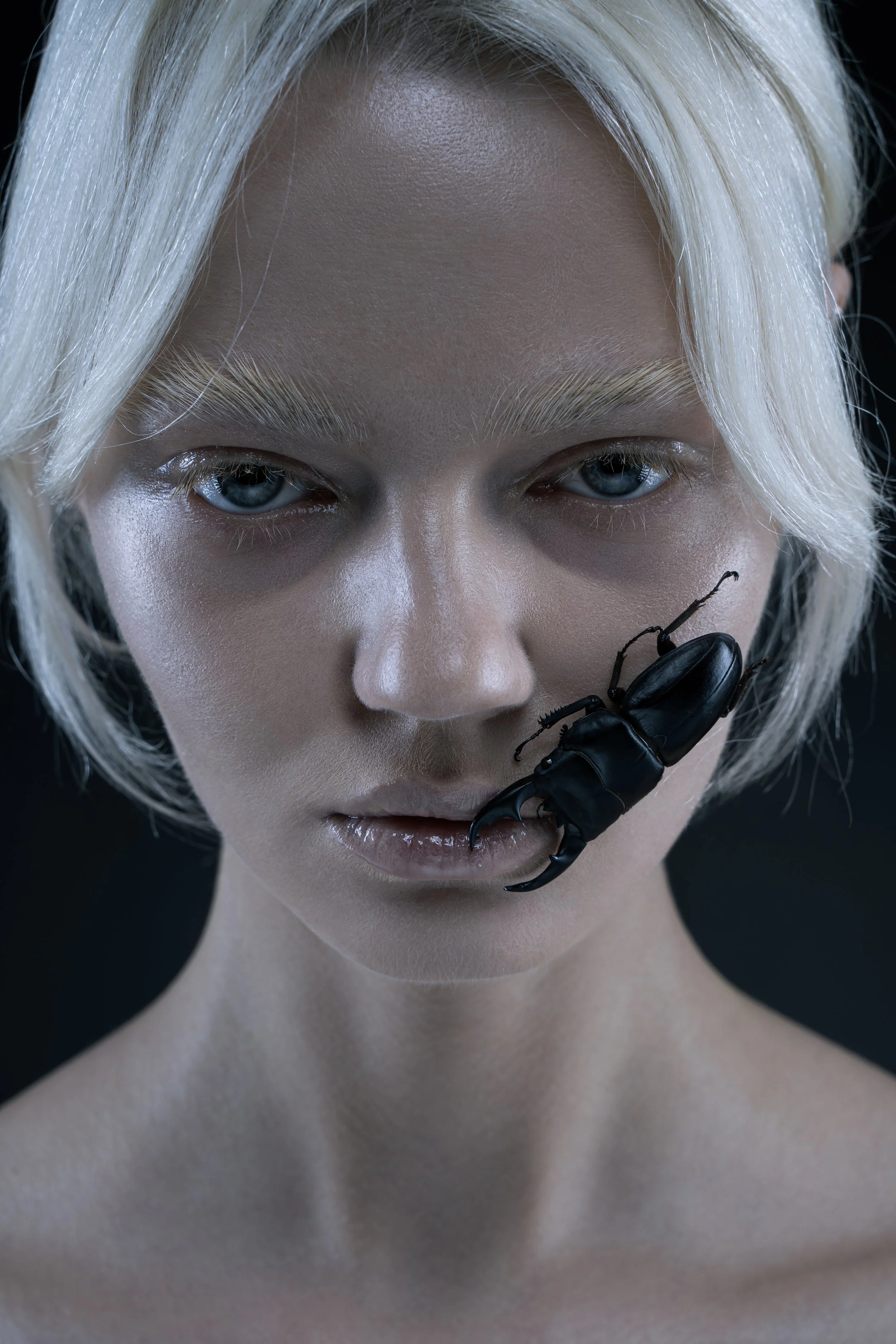 A woman with short, platinum blonde hair and light skin has a black beetle on her lips.