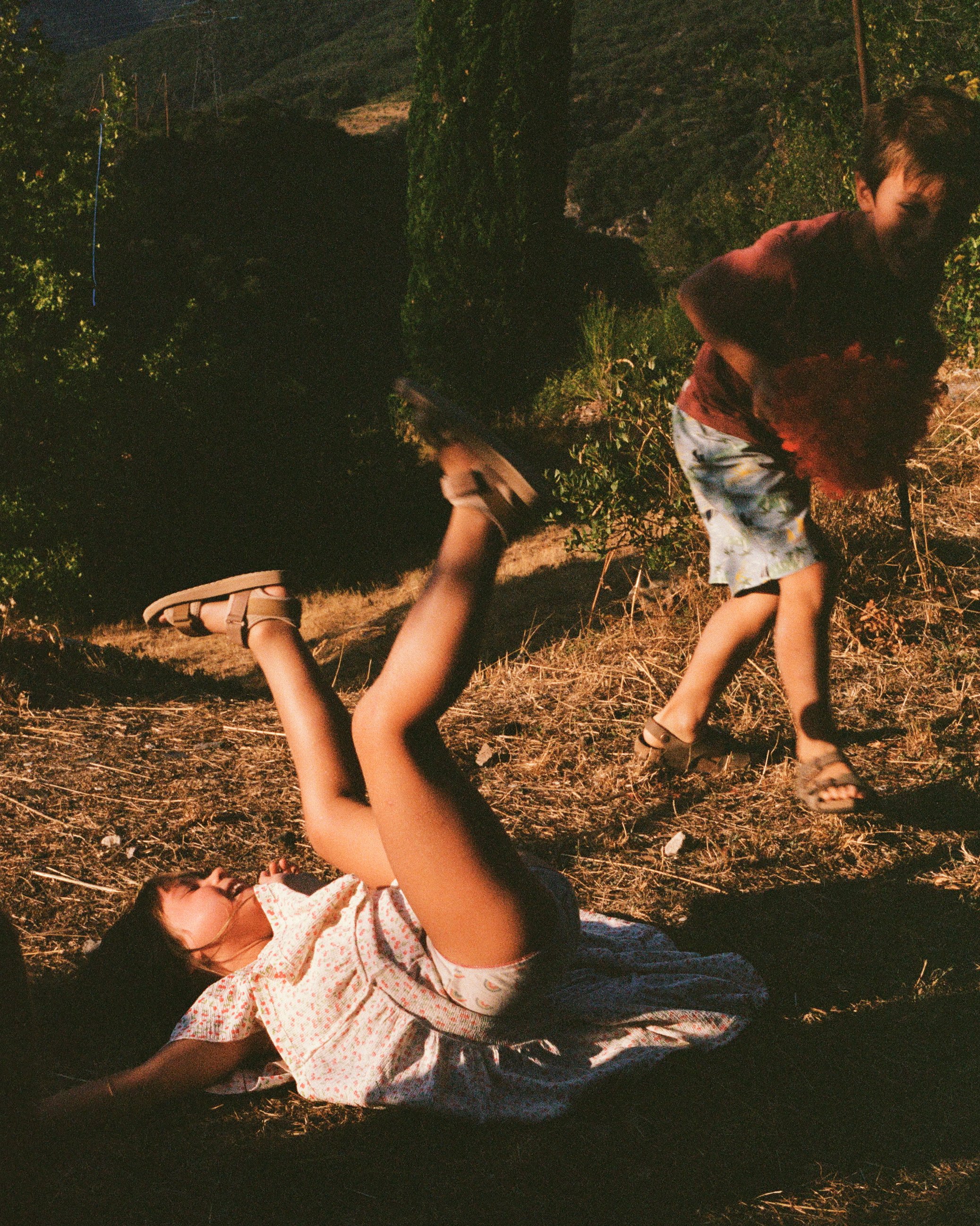Two children playing outdoors during daytime; one girl lying on the ground with her legs up, and one boy running with a ball near bushes and trees.