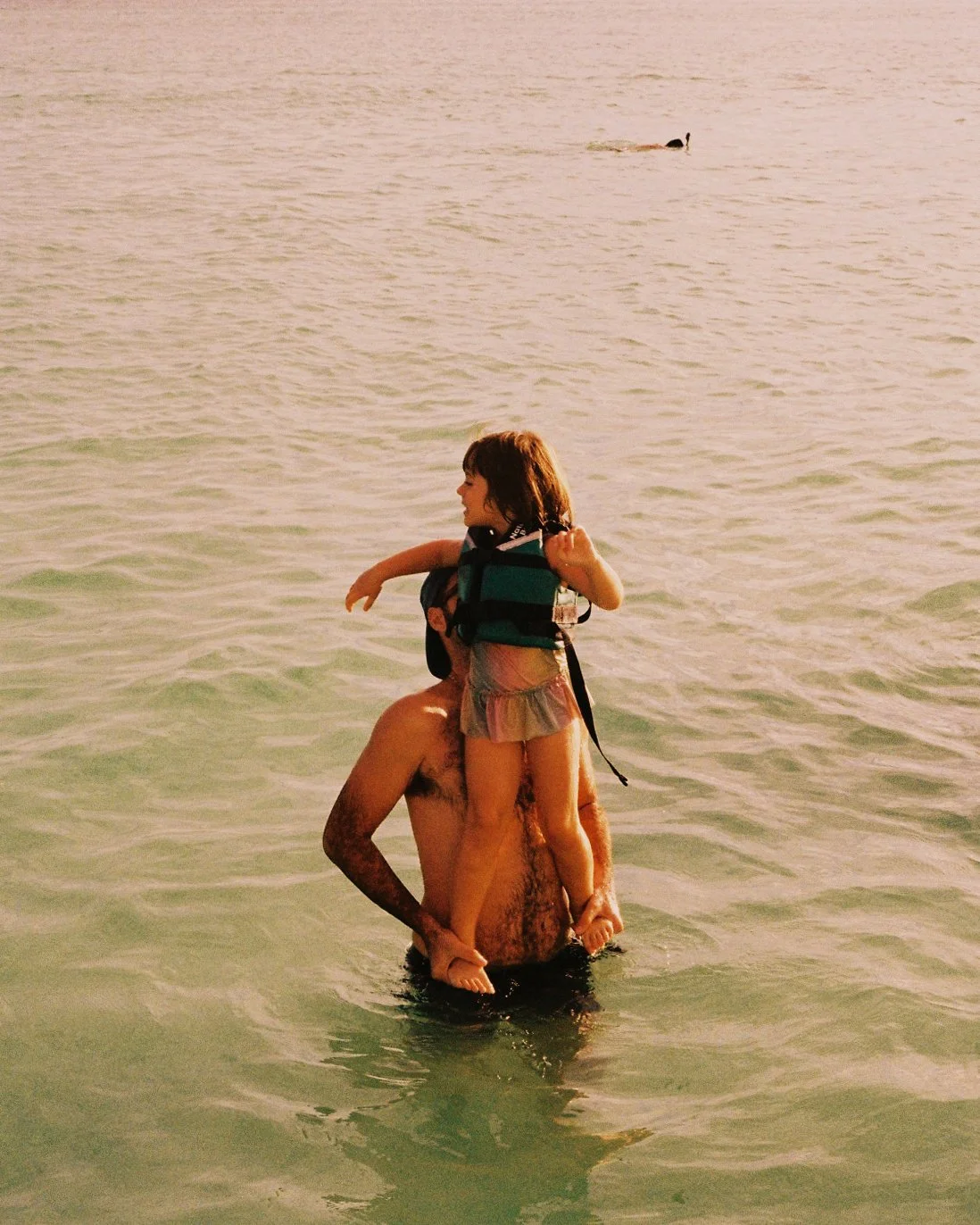 A man is holding a child on his shoulders in the ocean, with a dog swimming in the water in the background during sunset.