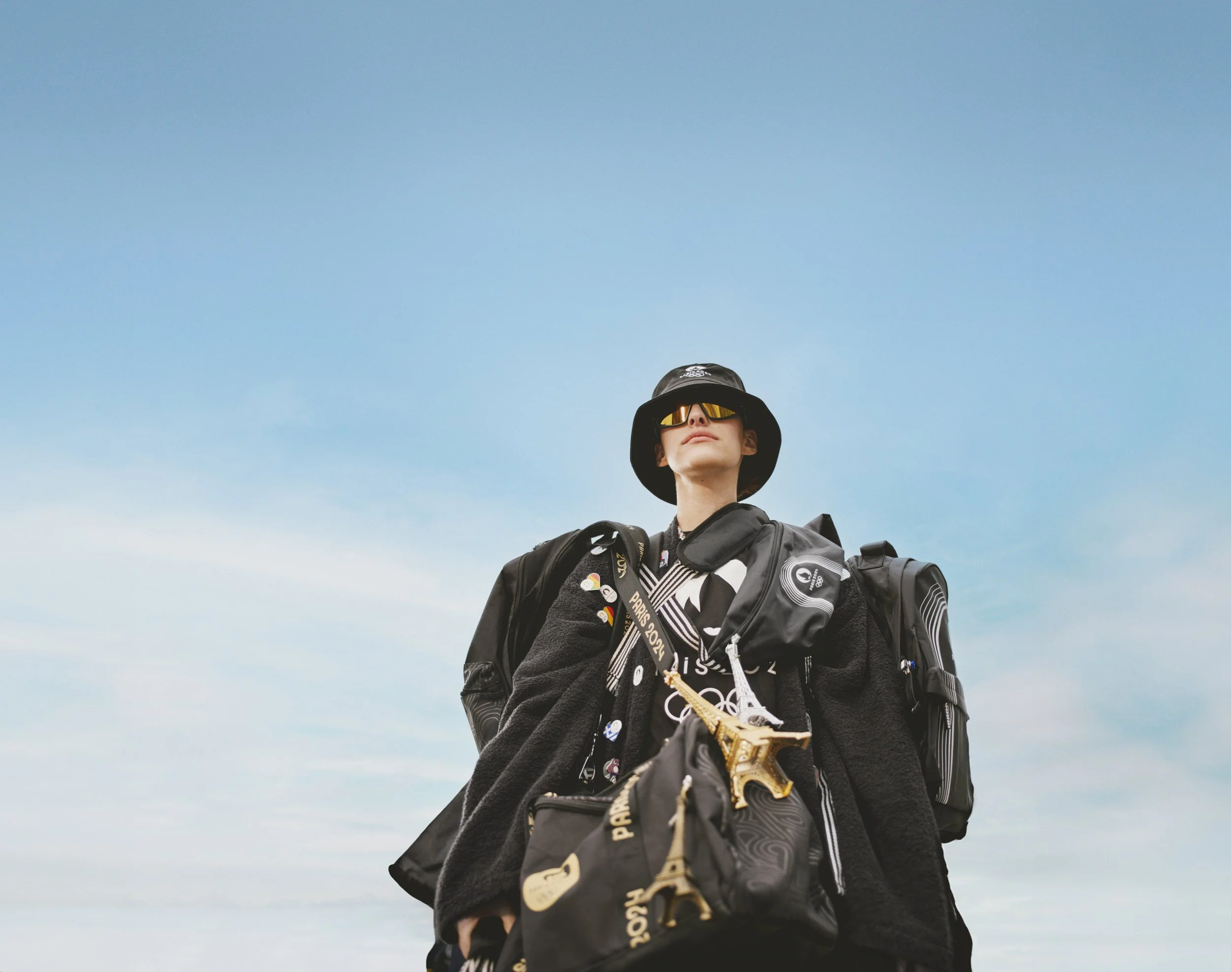 A woman wearing sunglasses, a black hat, and a black jacket, carrying multiple black bags and a gold Eiffel Tower keychain, standing outdoors against a blue sky.