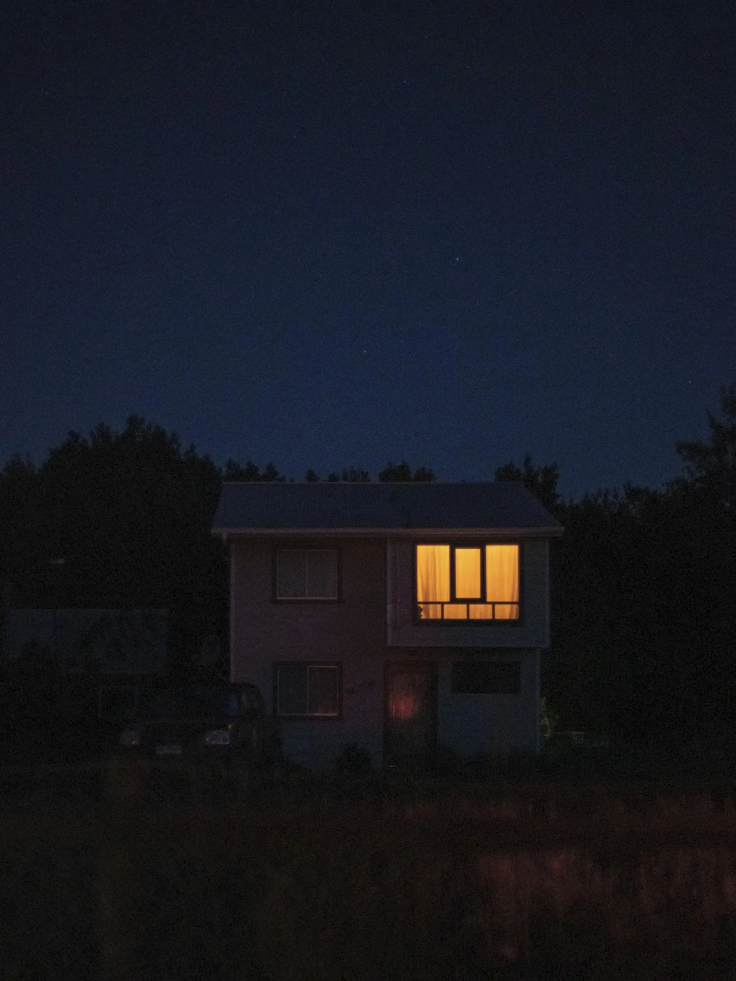 A house at night with a single illuminated window on the second floor, dark surroundings, and a starry sky.