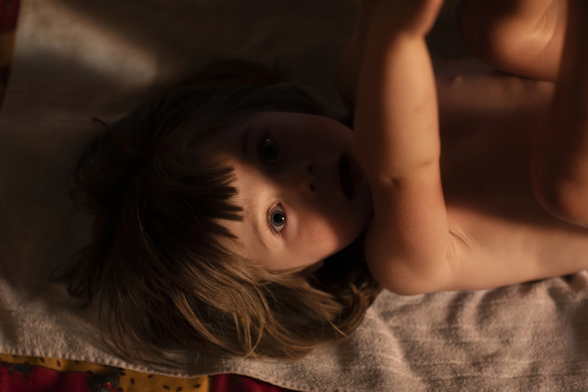 A young child laying on a soft surface, looking up with wide eyes, with light reflecting off their face.