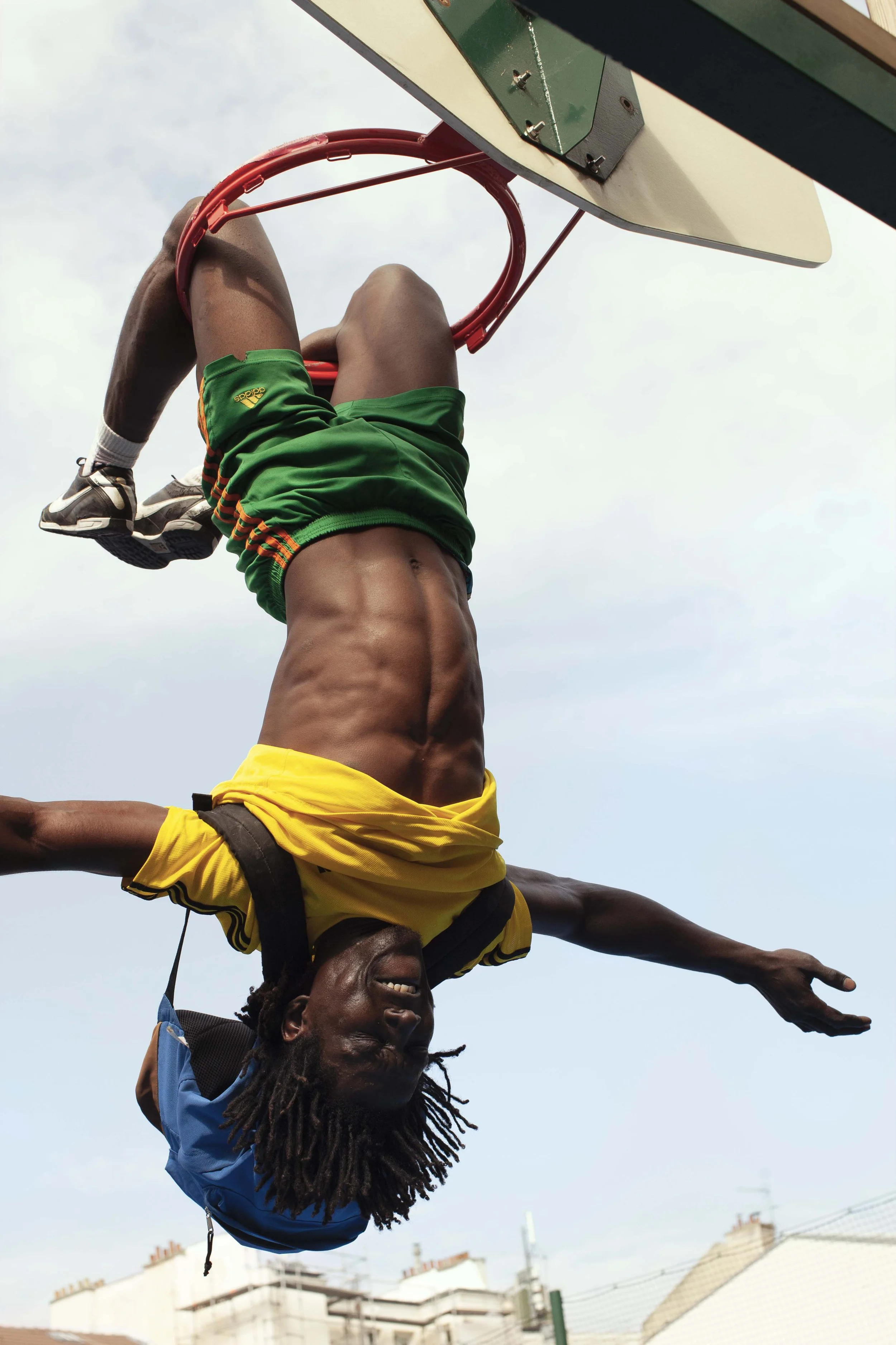 A man hanging upside down from a basketball hoop with one arm extended, smiling, wearing a yellow and black shirt, green shorts, and a blue cap.