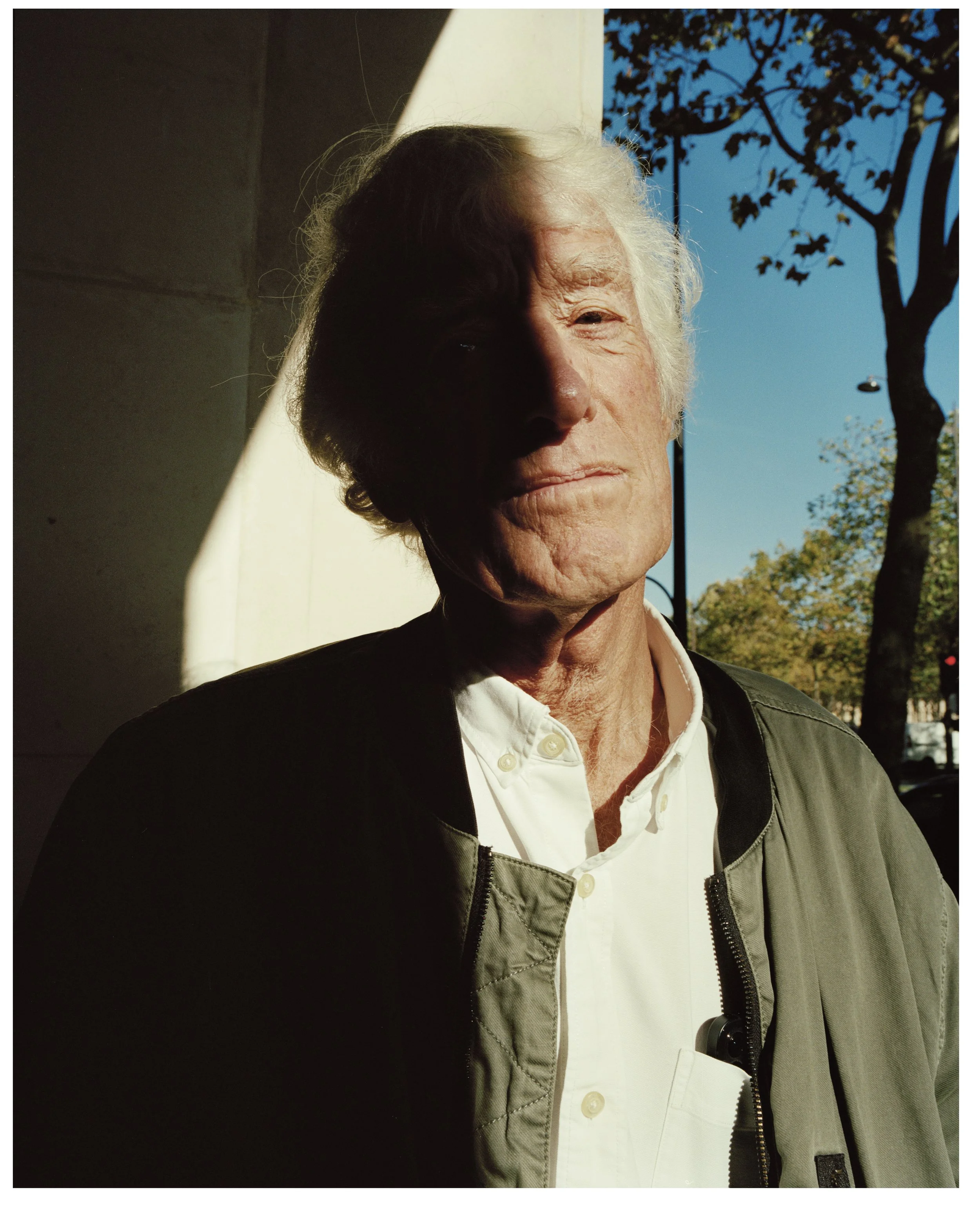A close-up of an elderly man with white hair, wearing a white collared shirt and a dark jacket, standing outside in sunlight, with a tree and blue sky in the background.