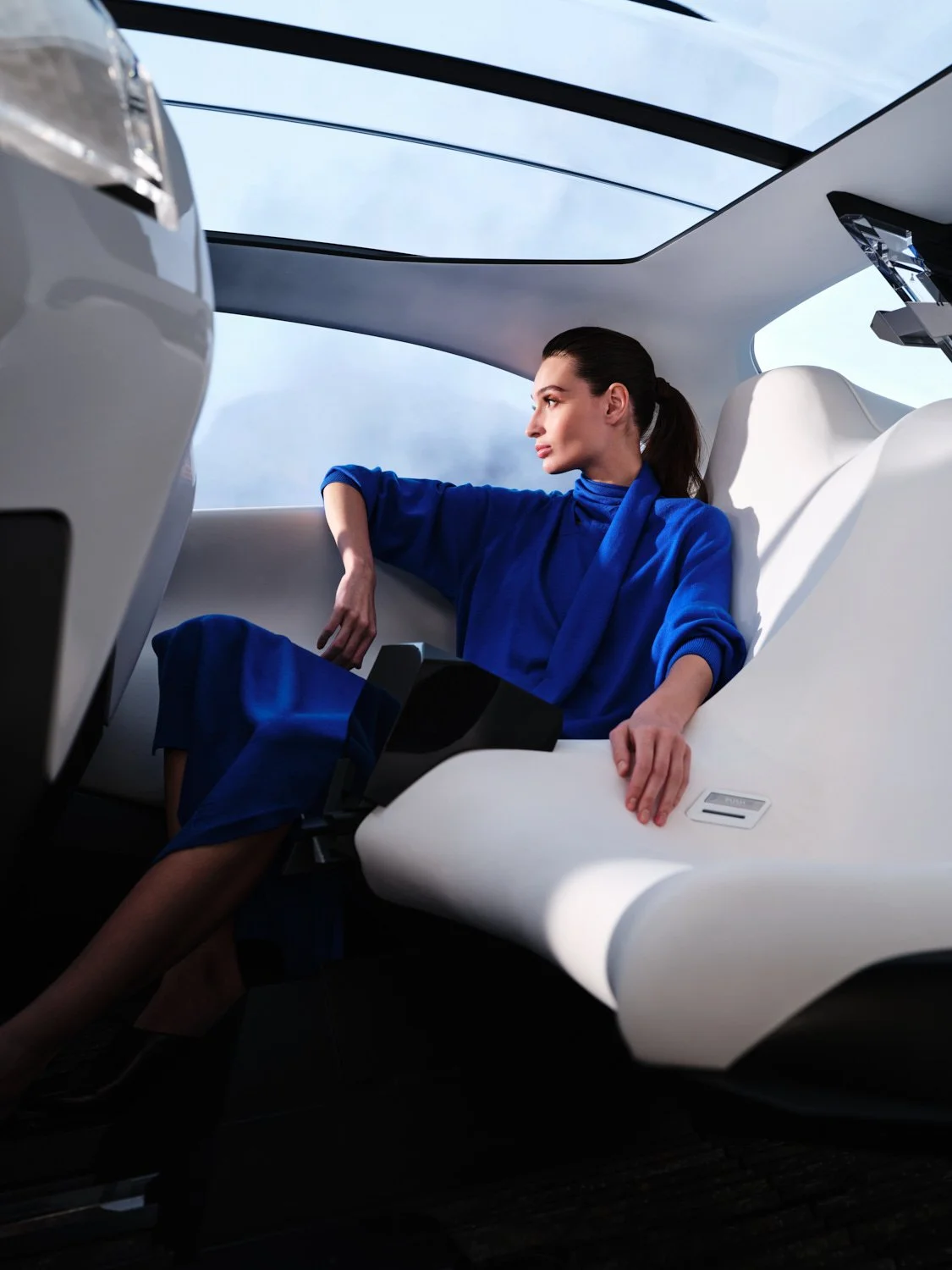 A woman in a blue outfit sitting in a modern, white, high-tech vehicle with a panoramic glass roof, looking out at the sky.