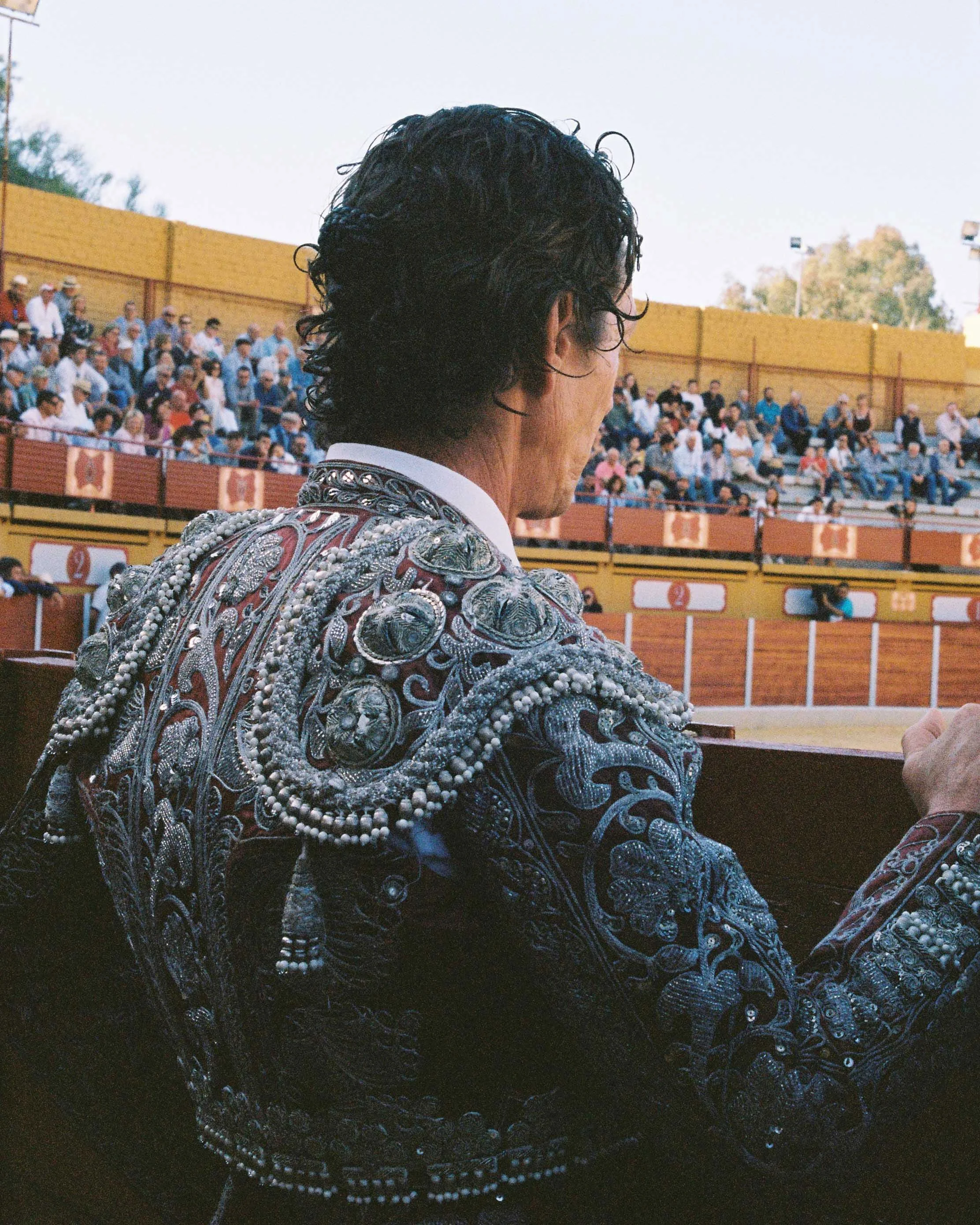 Bullfighter wearing a traditional ornate jacket with embroidery and beads in a bullfighting arena with spectators in the background.