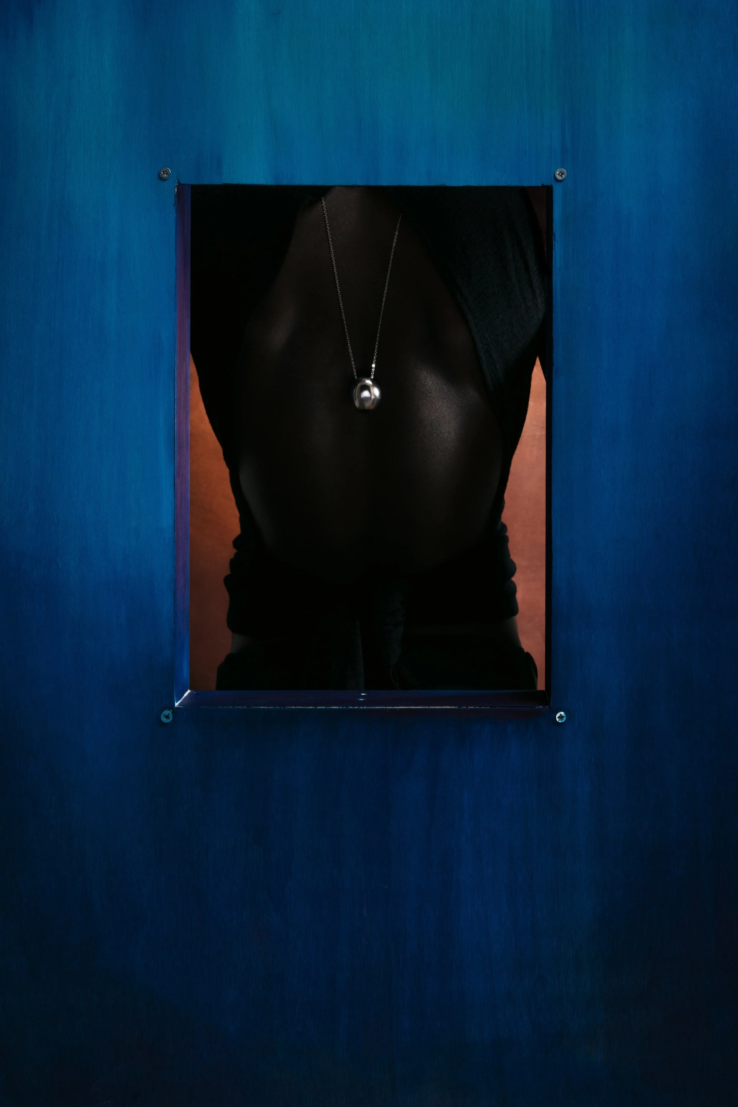 Person wearing a black shirt with a silver pendant necklace, seen through a small rectangular opening in a blue wall.