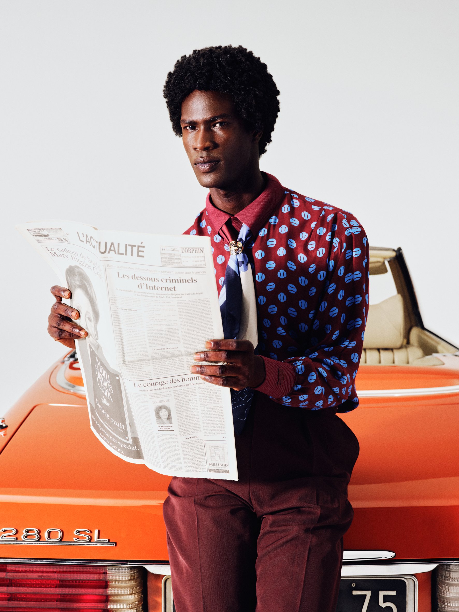 A man with dark skin, curly black hair, wearing a patterned maroon shirt with blue spots, and a patterned tie, stands in front of an orange vintage car, holding a newspaper.