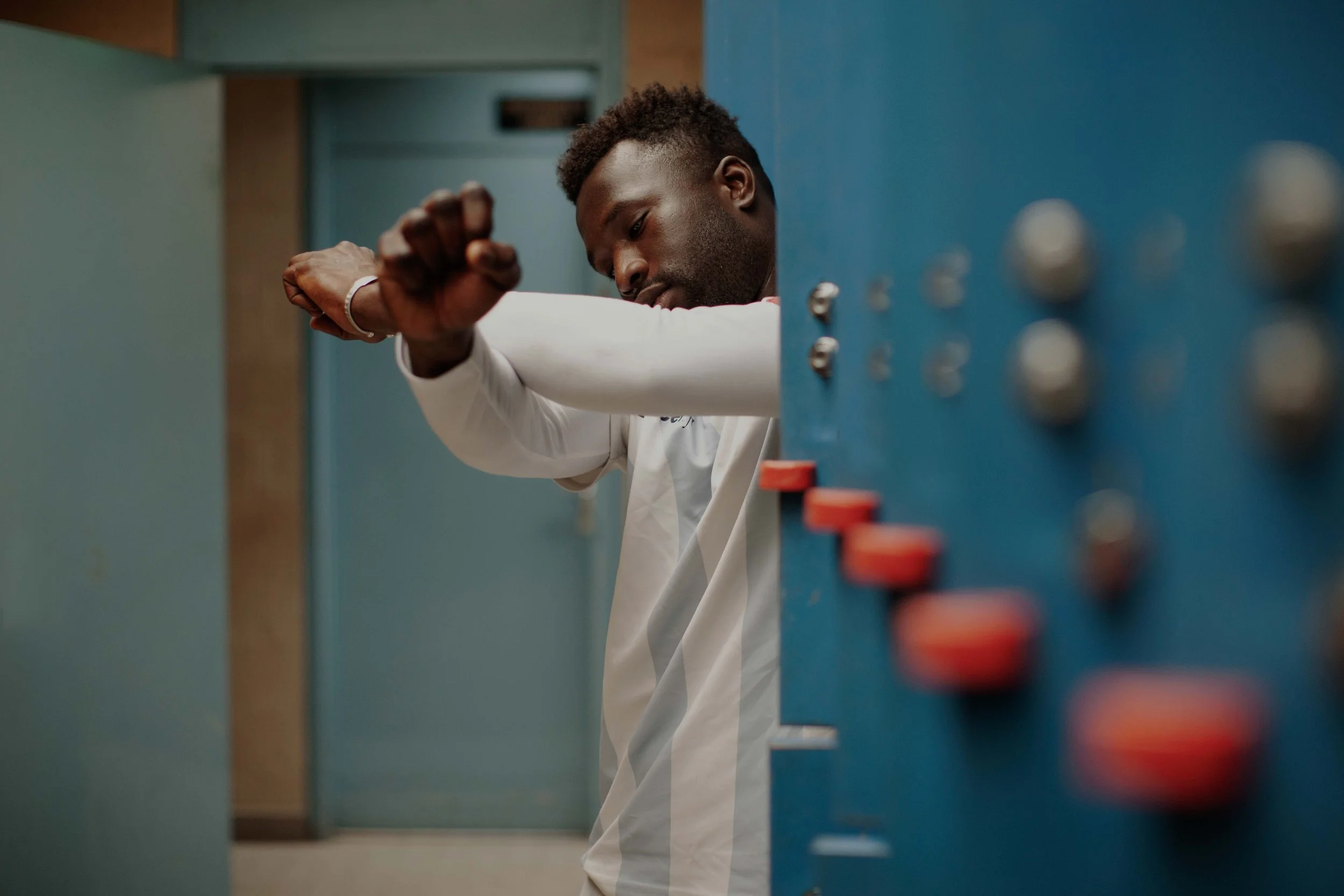 A man in a white shirt is checking his watch in a hallway near a blue panel with buttons or switches.