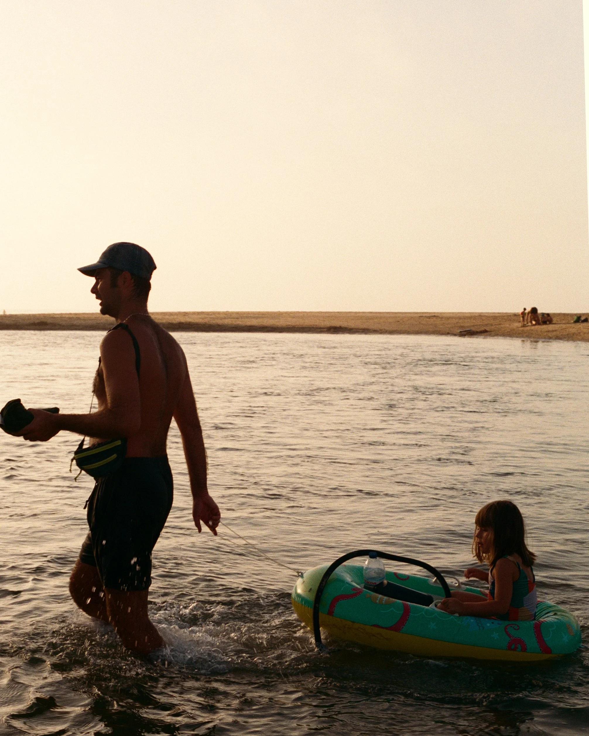 A man and a young girl in a flotation device, at a beach or lake, during sunset or sunrise.
