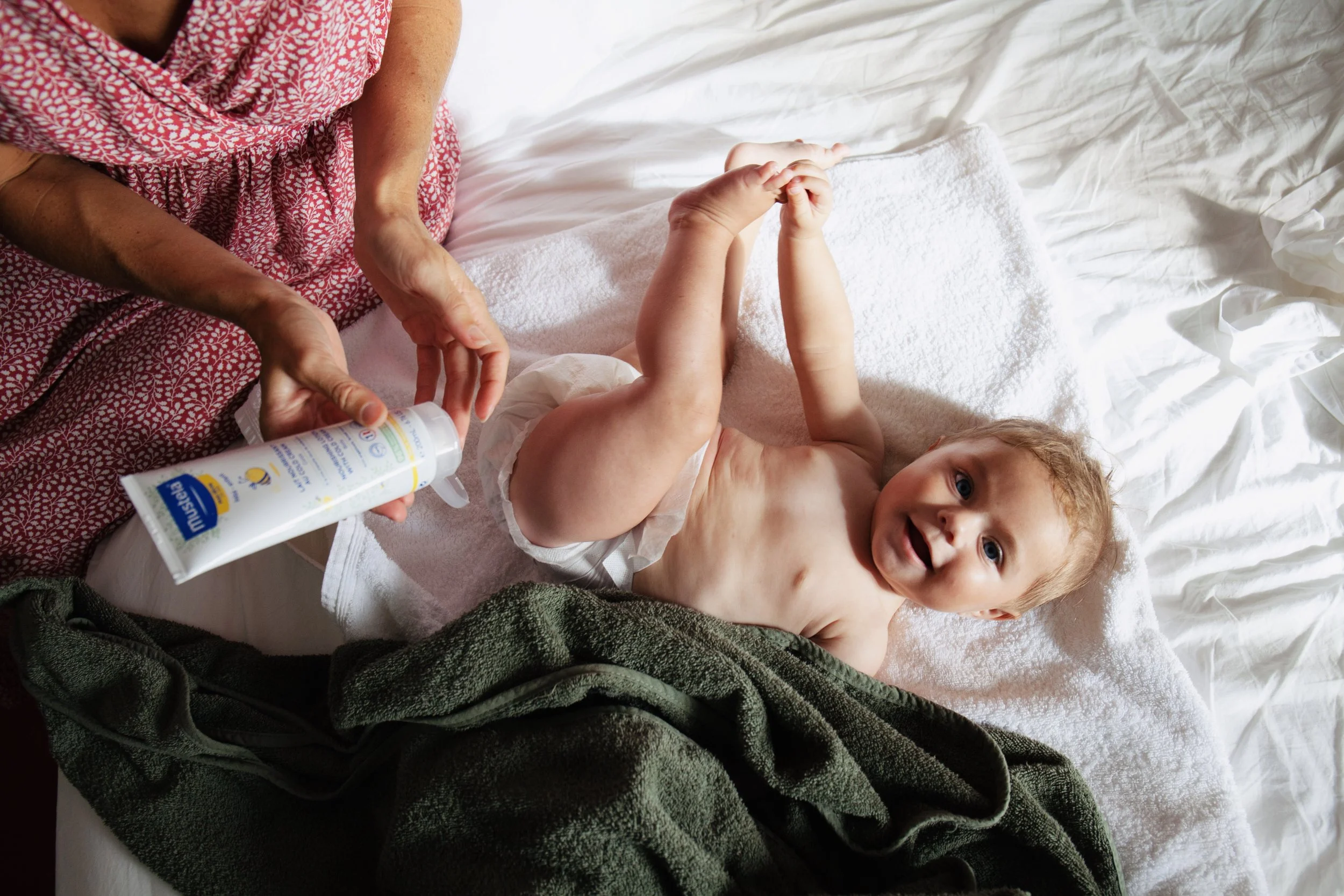 A baby lying on a bed, smiling and looking at the camera. An adult is holding a bottle of baby lotion or sunscreen, preparing to apply it on the baby. The baby is partially covered with a green towel and the bed has white sheets and pillowcases.