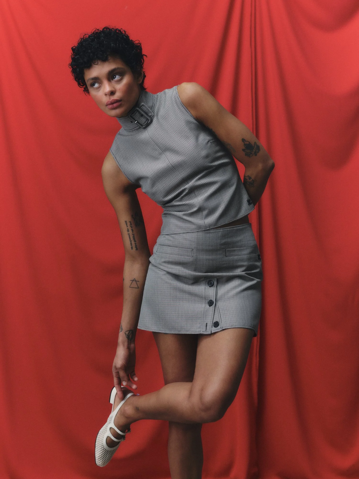 A woman with short curly hair wearing a grey sleeveless checkered dress, white sneakers, standing against a red fabric background.