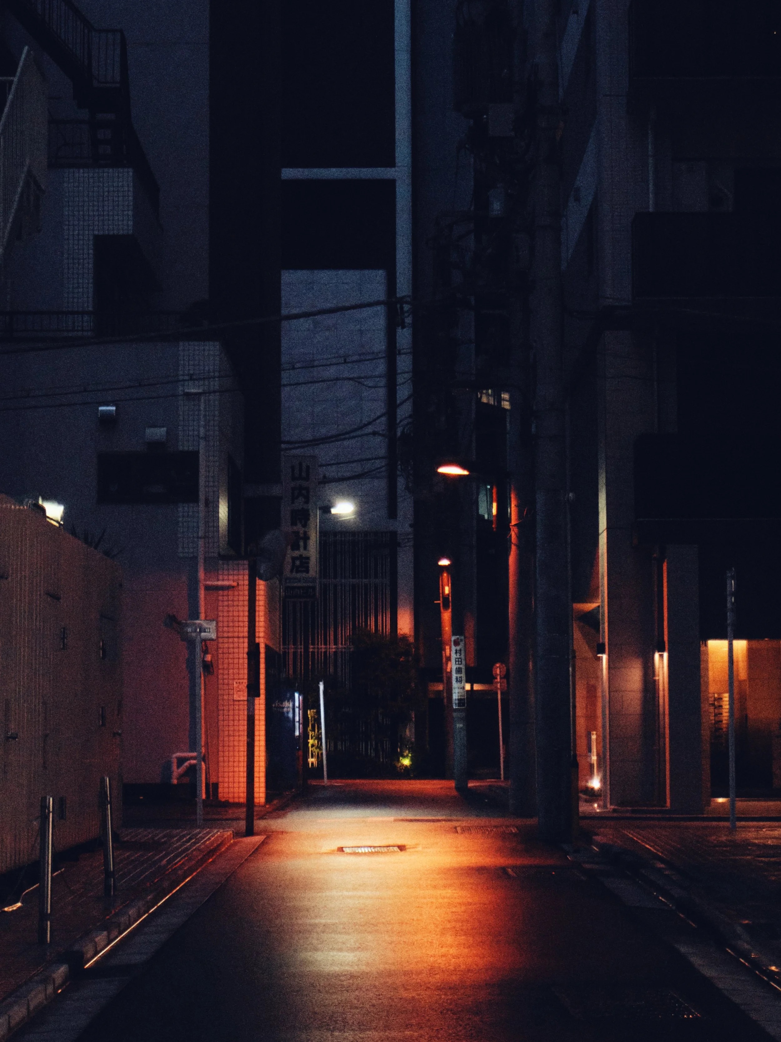 Nighttime city street with dim streetlights, utility poles, and buildings, some with illuminated signs, in an urban area.