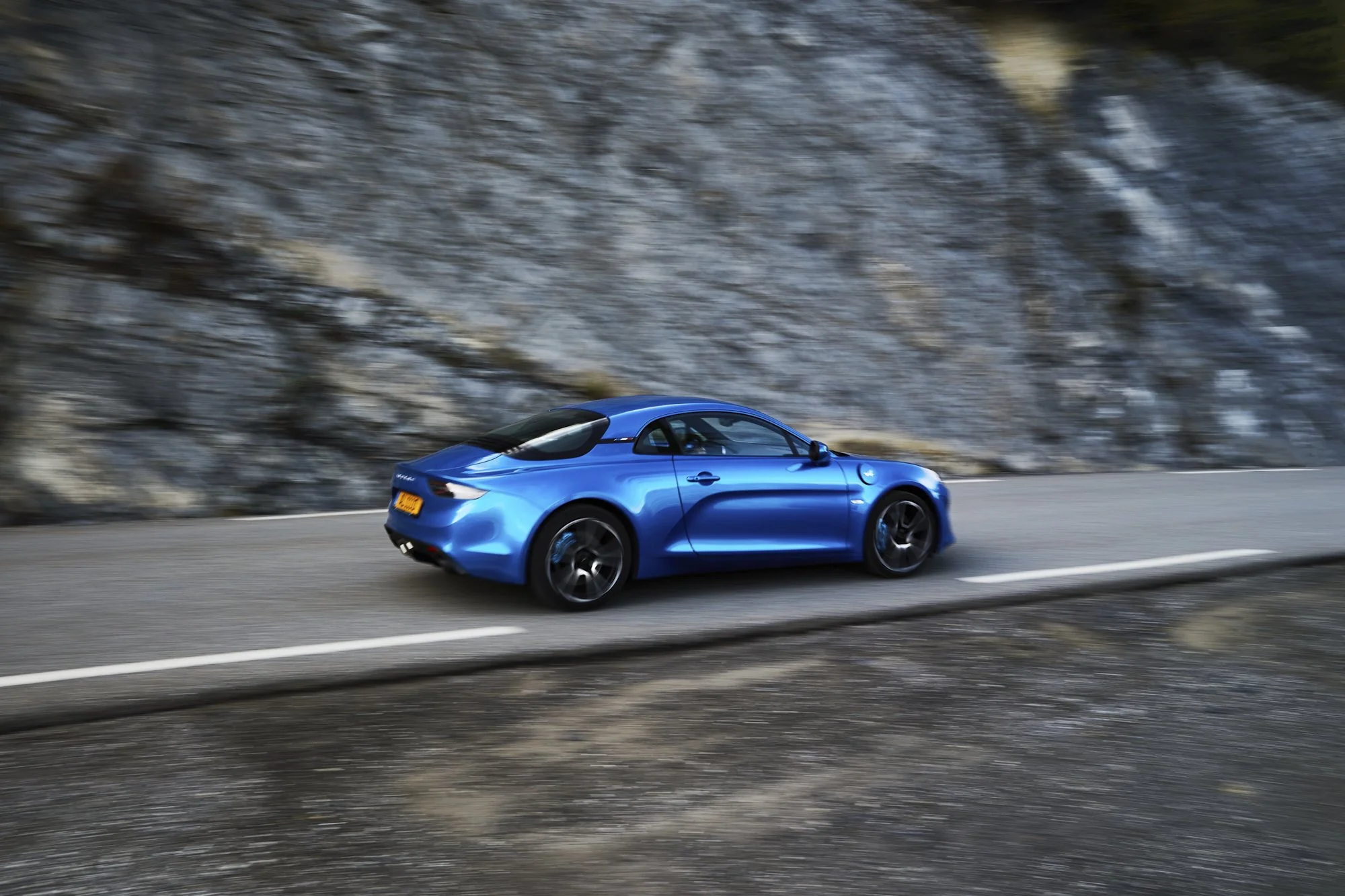 A blue sports car driving on a mountain road with rocky cliffside in the background.