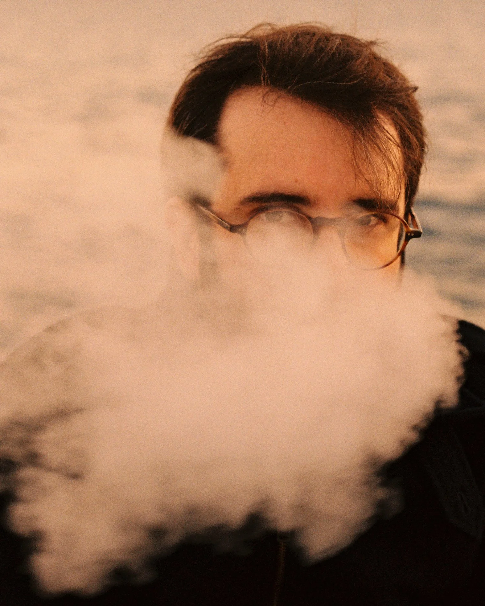 A person with dark hair and glasses is partially obscured by vapor or smoke, with water in the background.