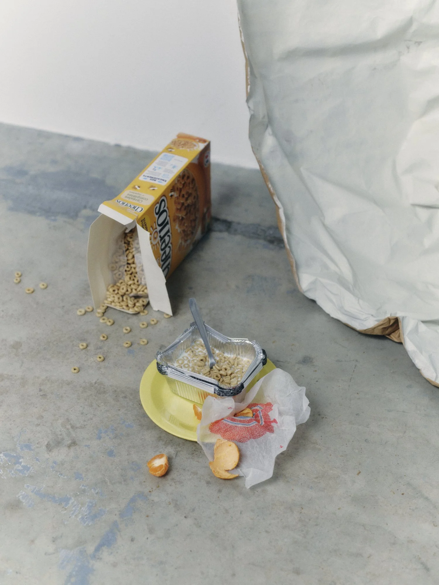 An overturned cereal box spilled dry cereal onto the floor next to an open container of cereal and a yellow plate. Food wrappers and broken orange slices are scattered on the floor.