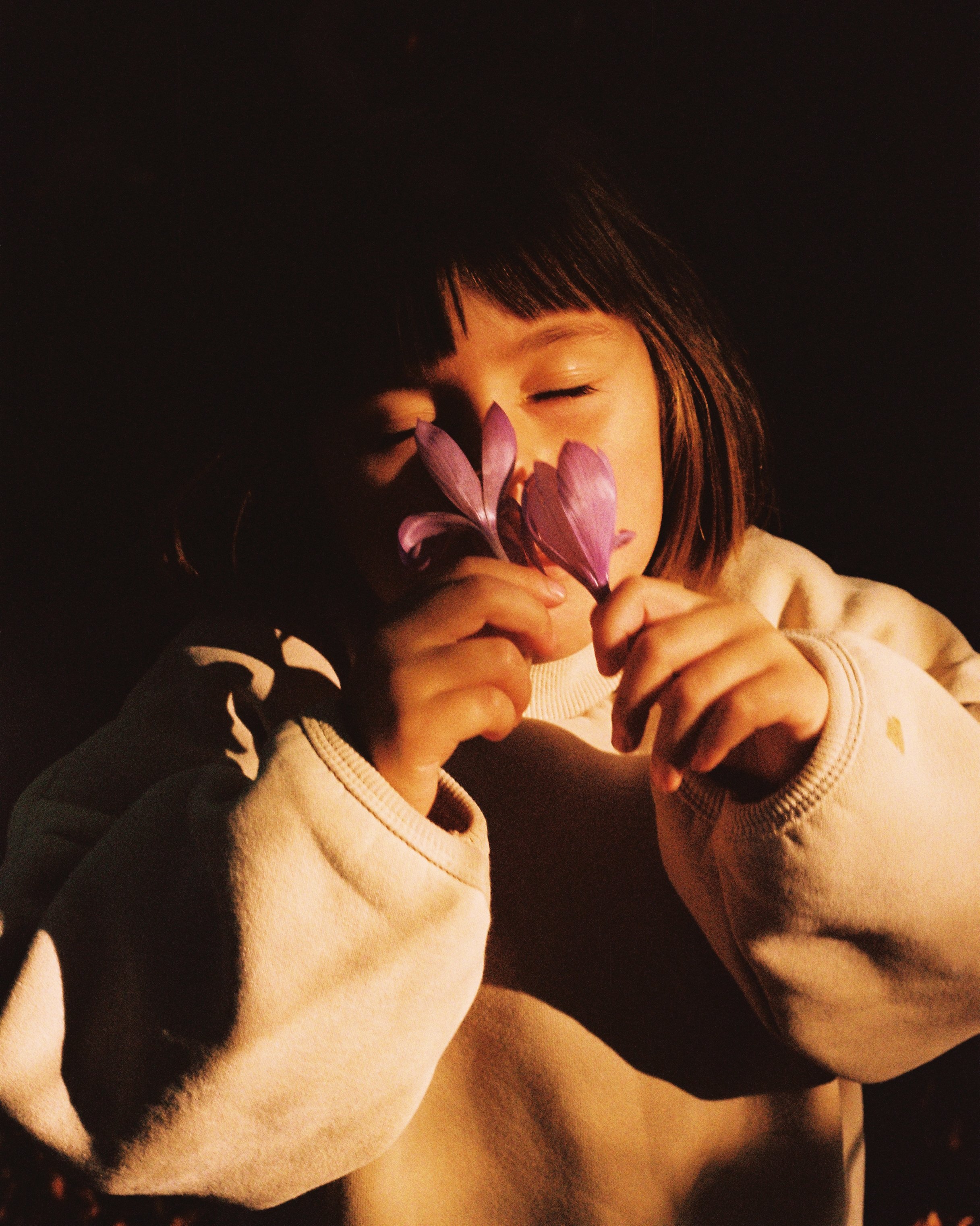 A young girl with short brown hair and a beige sweatshirt is holding purple flowers close to her face with her eyes closed in a dark setting.