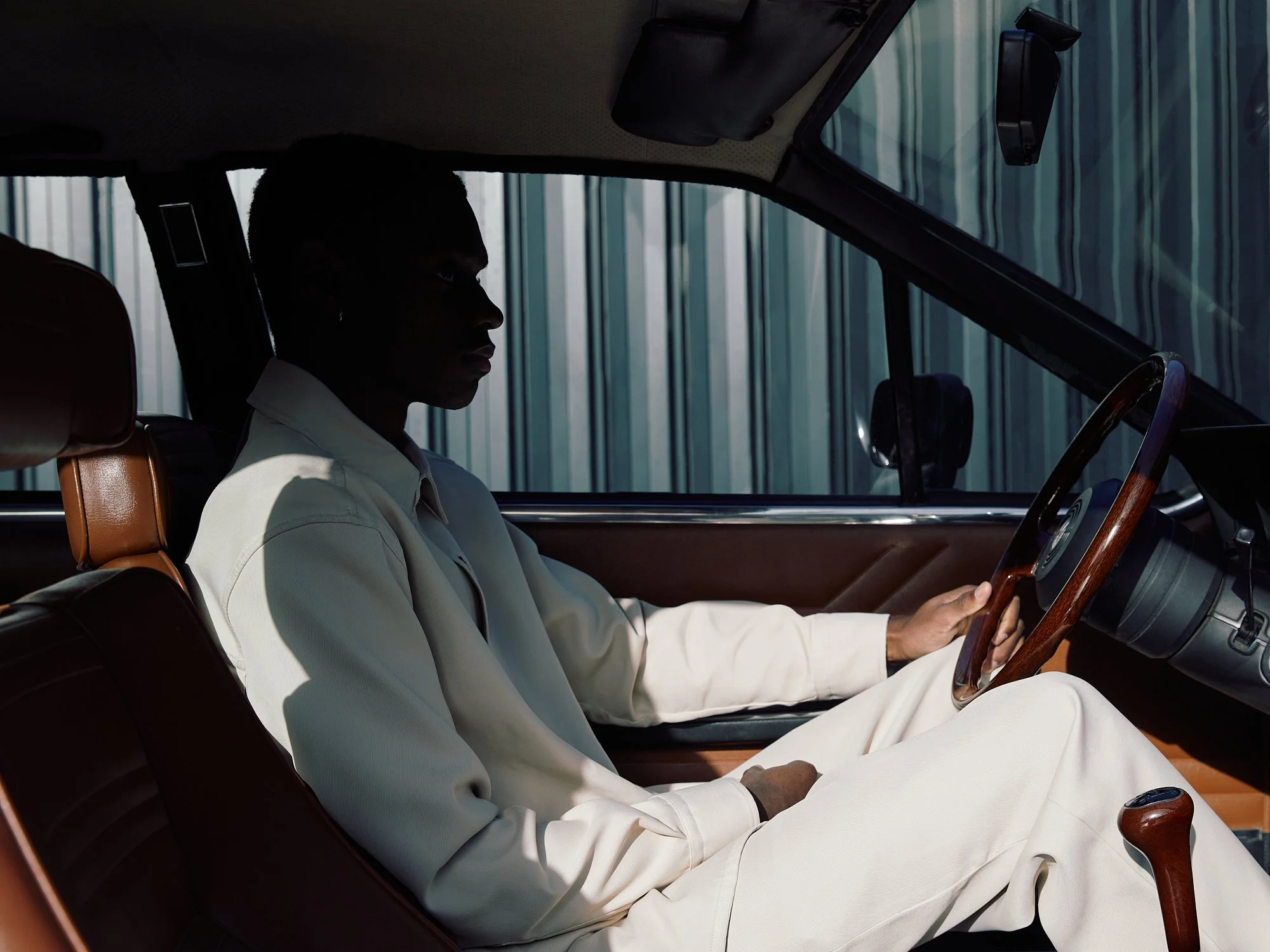 Person with dark skin sitting in the driver's seat of a vintage car, holding the steering wheel. The person is wearing a light-colored outfit. The car's interior features brown leather seats and a wooden steering wheel. The background shows a metallic fence or wall outside the car window.