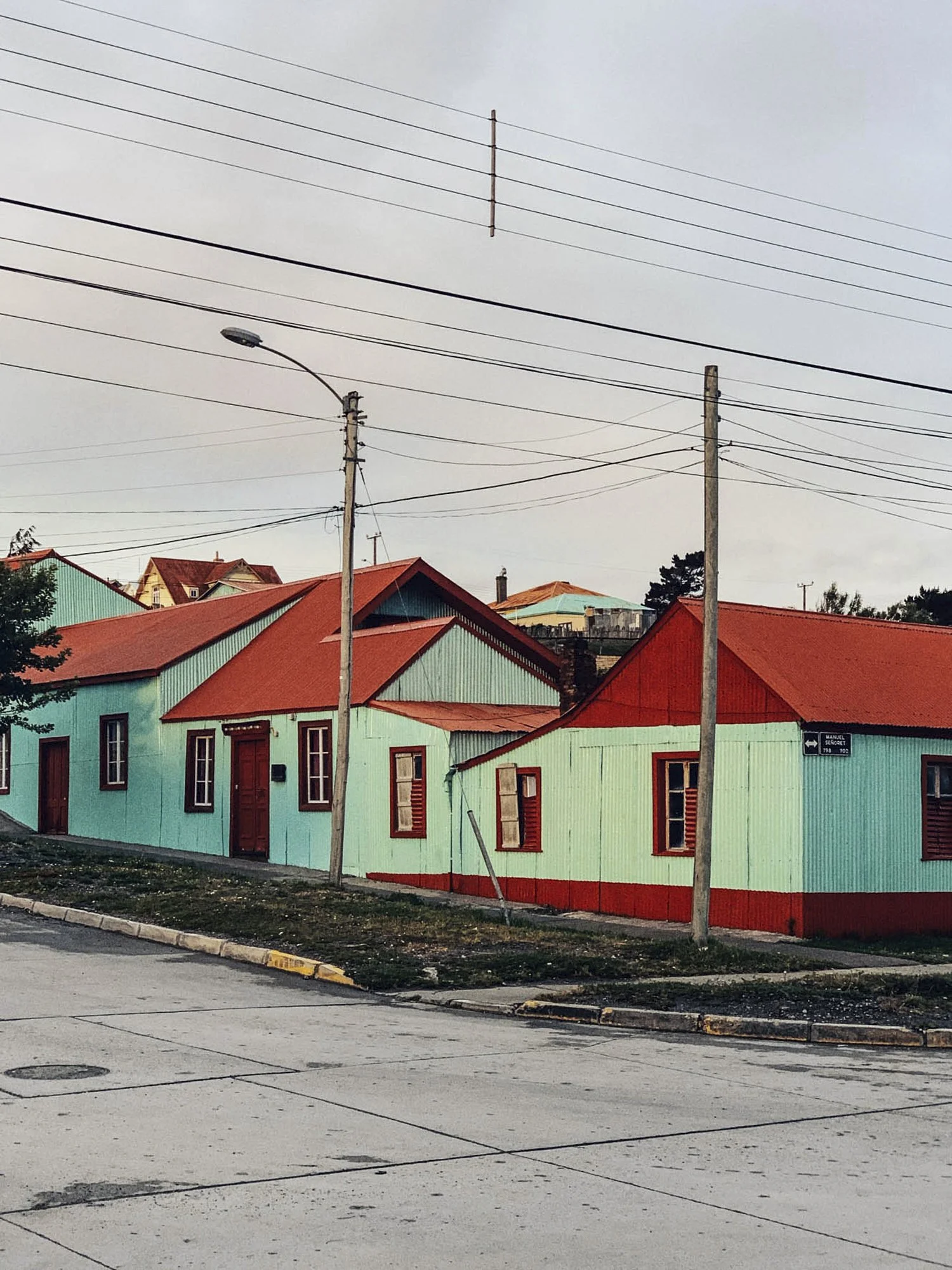 A colorful street scene with pastel green houses featuring red roofs, a sidewalk, utility poles, and power lines, under a gray sky.