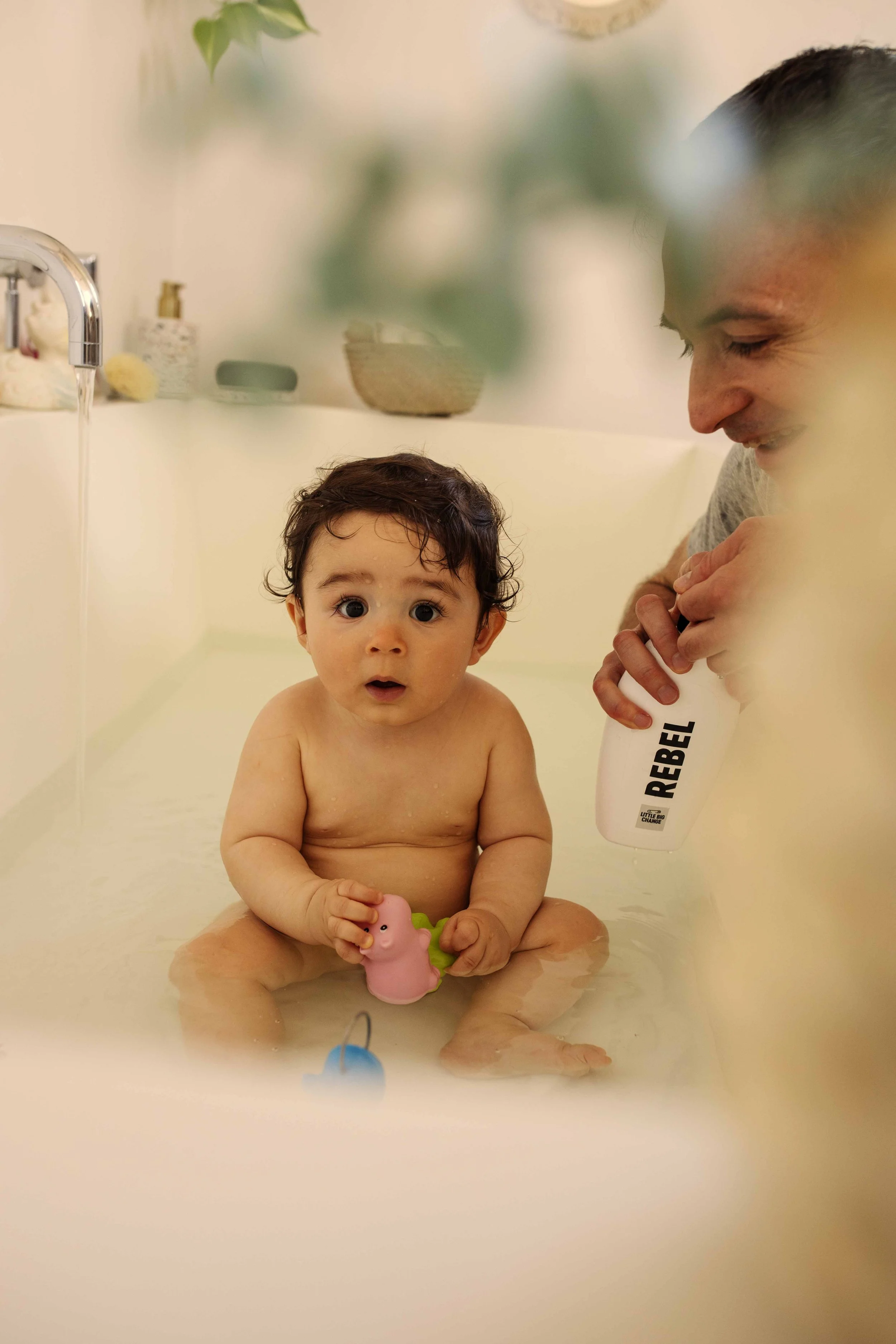 A toddler sitting in a bathtub with a surprised expression, holding a pink and green bath toy, while an adult nearby holds a bottle labeled "REBEL" and is smiling.