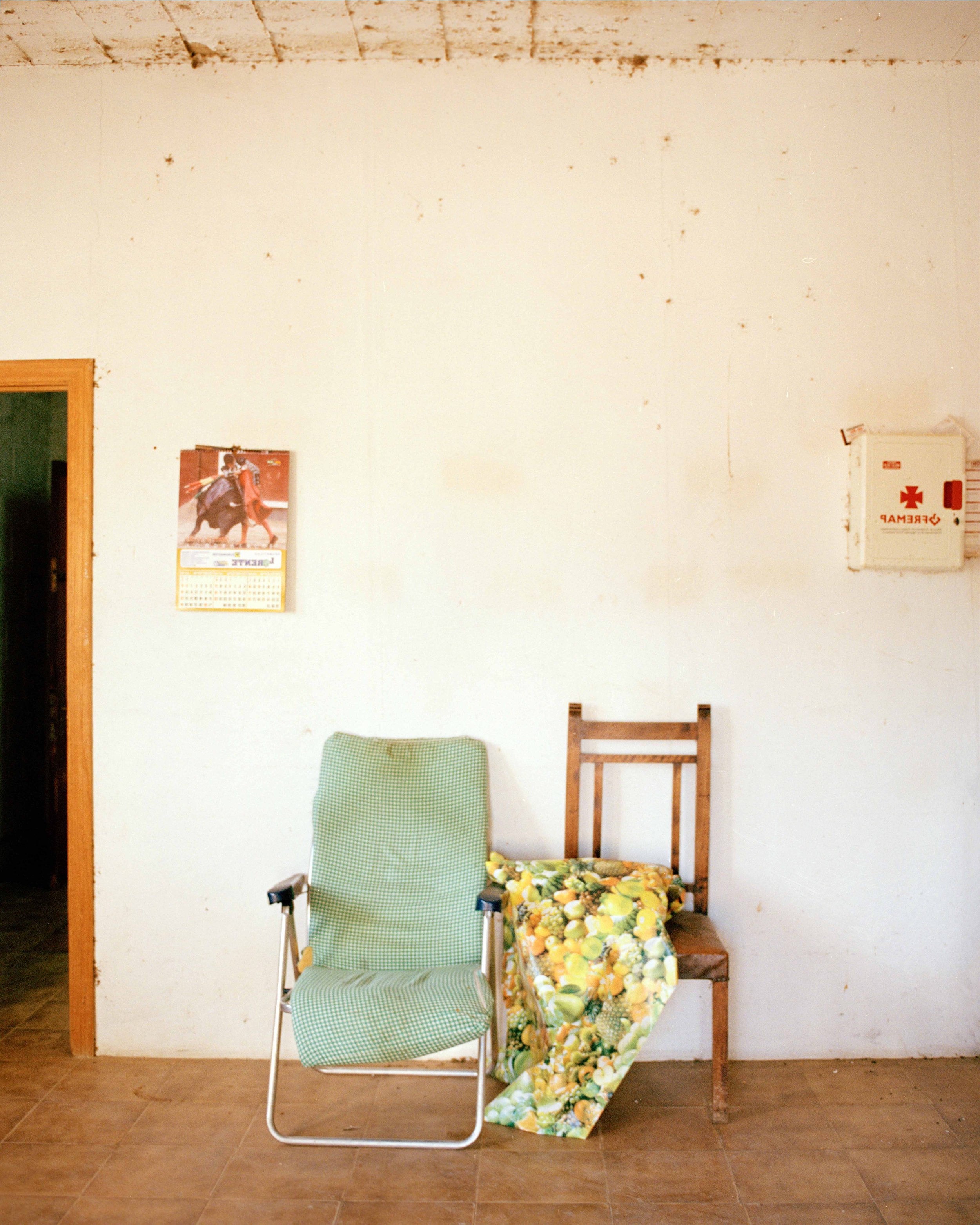 A plain white wall with two chairs: one green armchair and one wooden chair with a floral cloth. A calendar with a horse on it hangs on the wall. An emergency box is mounted on the wall.