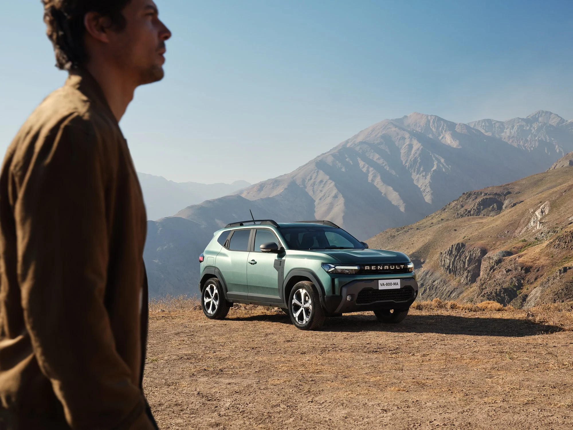 A man stands in the foreground with a mountainous landscape and a green Renault car in the background.