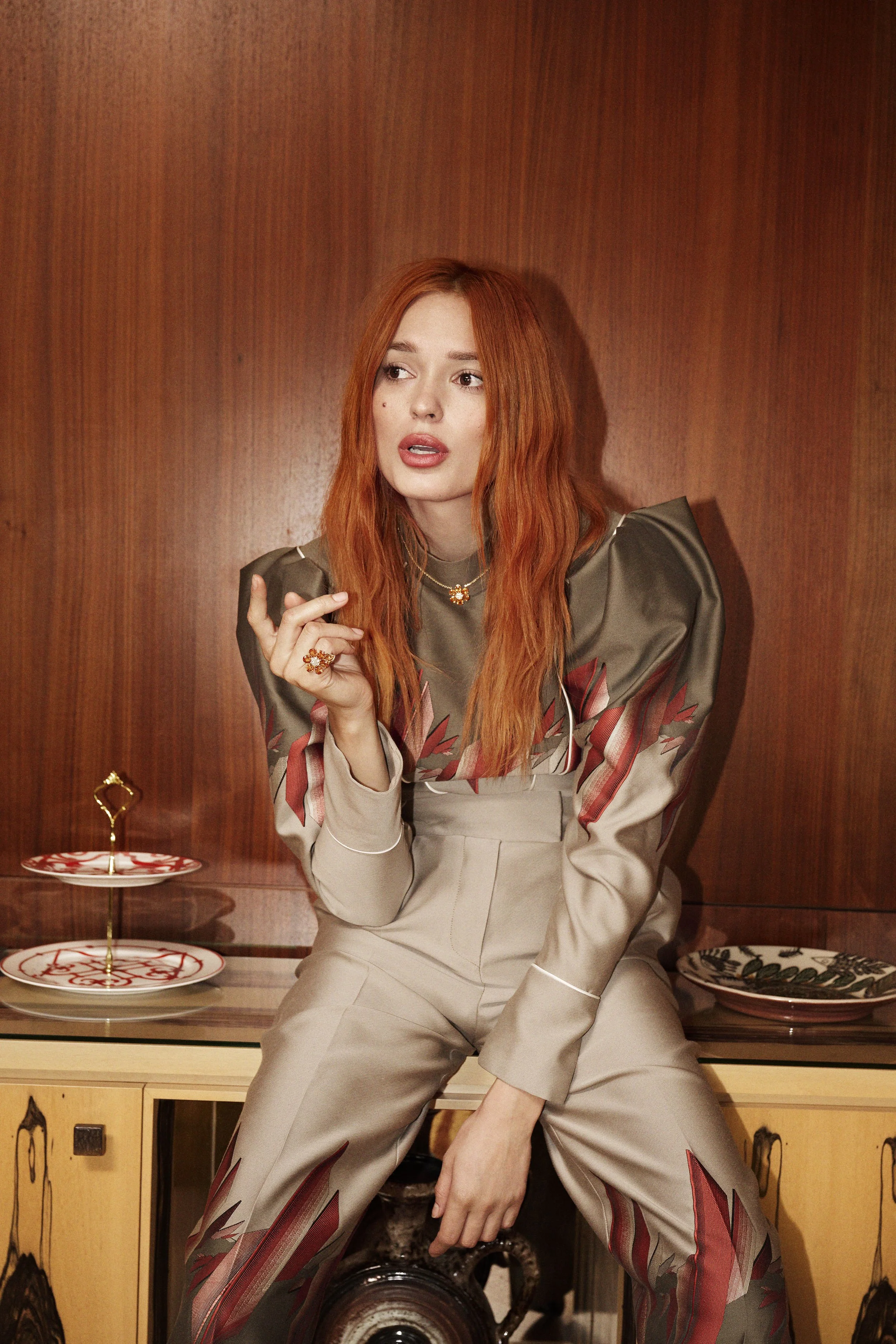 A woman with long red hair sitting on a wooden cabinet against a wood-paneled wall, dressed in a satin top and matching pants with red and green leaf patterns, gesturing with her right hand.