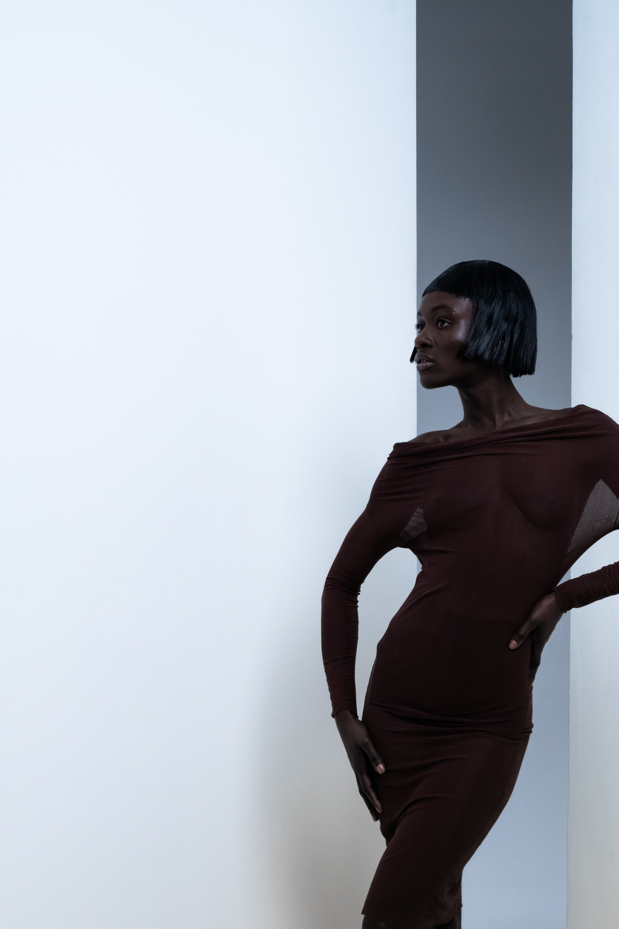 A woman with short black hair in a brown dress poses against a white and gray wall.