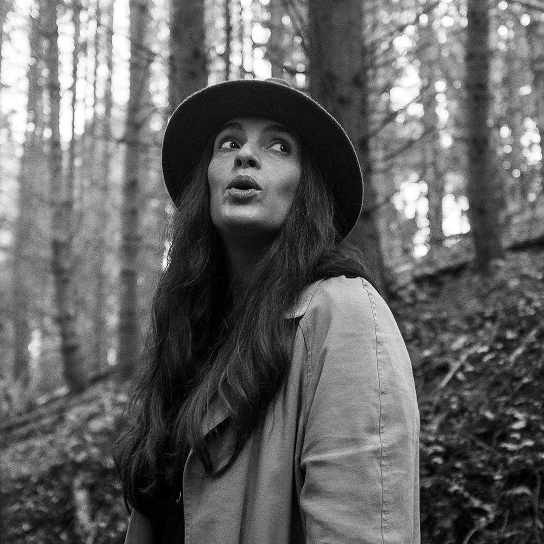 A woman with long wavy hair wearing a wide-brimmed hat and a jacket, standing outdoors in a forest with trees in the background.