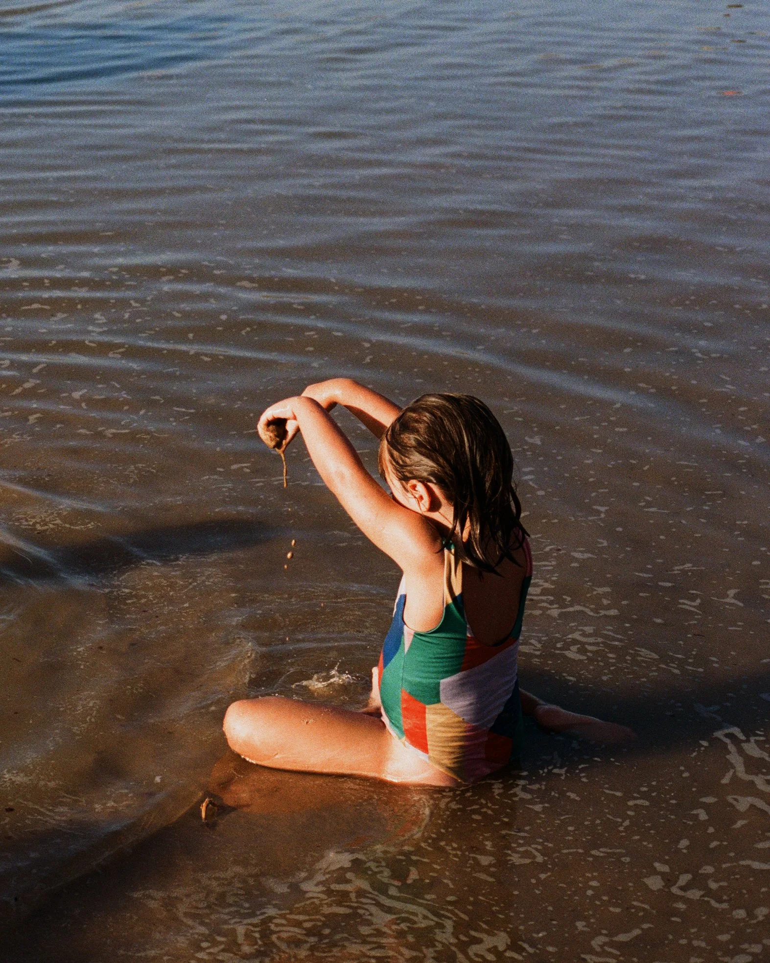 A young girl in a colorful swimsuit sitting in shallow water near the shore, playing with a stick.
