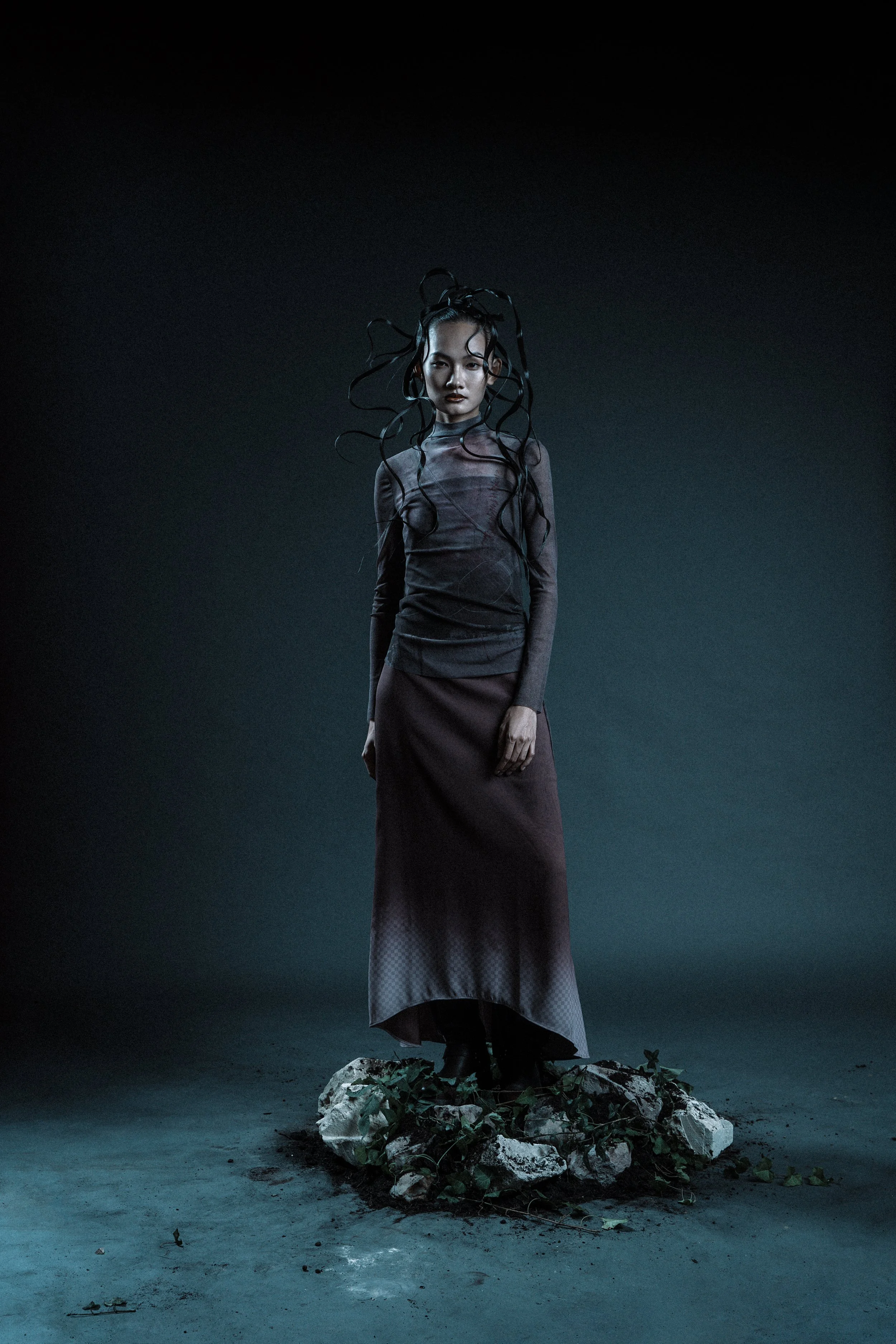 A woman with long, curly black hair standing on a stone and dirt platform, wearing a dark, long-sleeved top and a long skirt, in a dark, moody setting.
