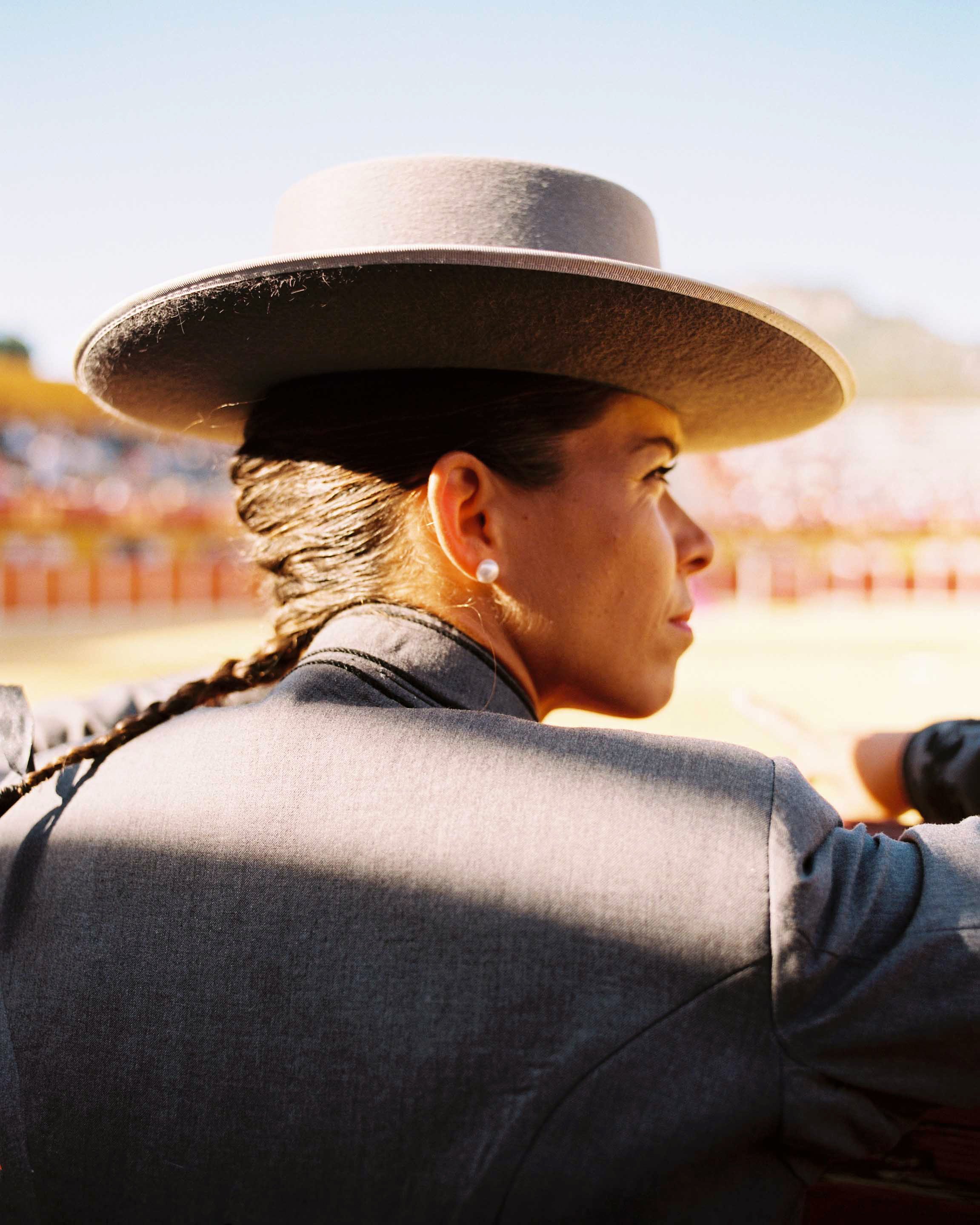 A woman wearing a large beige wide-brimmed hat, pearl earrings, a dark gray blazer, and black gloves, looking to the right outdoors during the daytime.