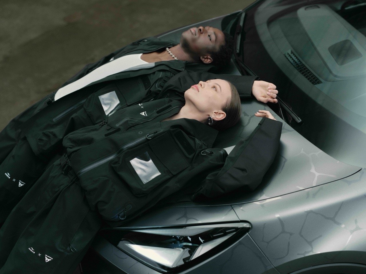 A man and woman lying on the hood of a gray sports car, eyes closed, relaxing with their arms stretched out.