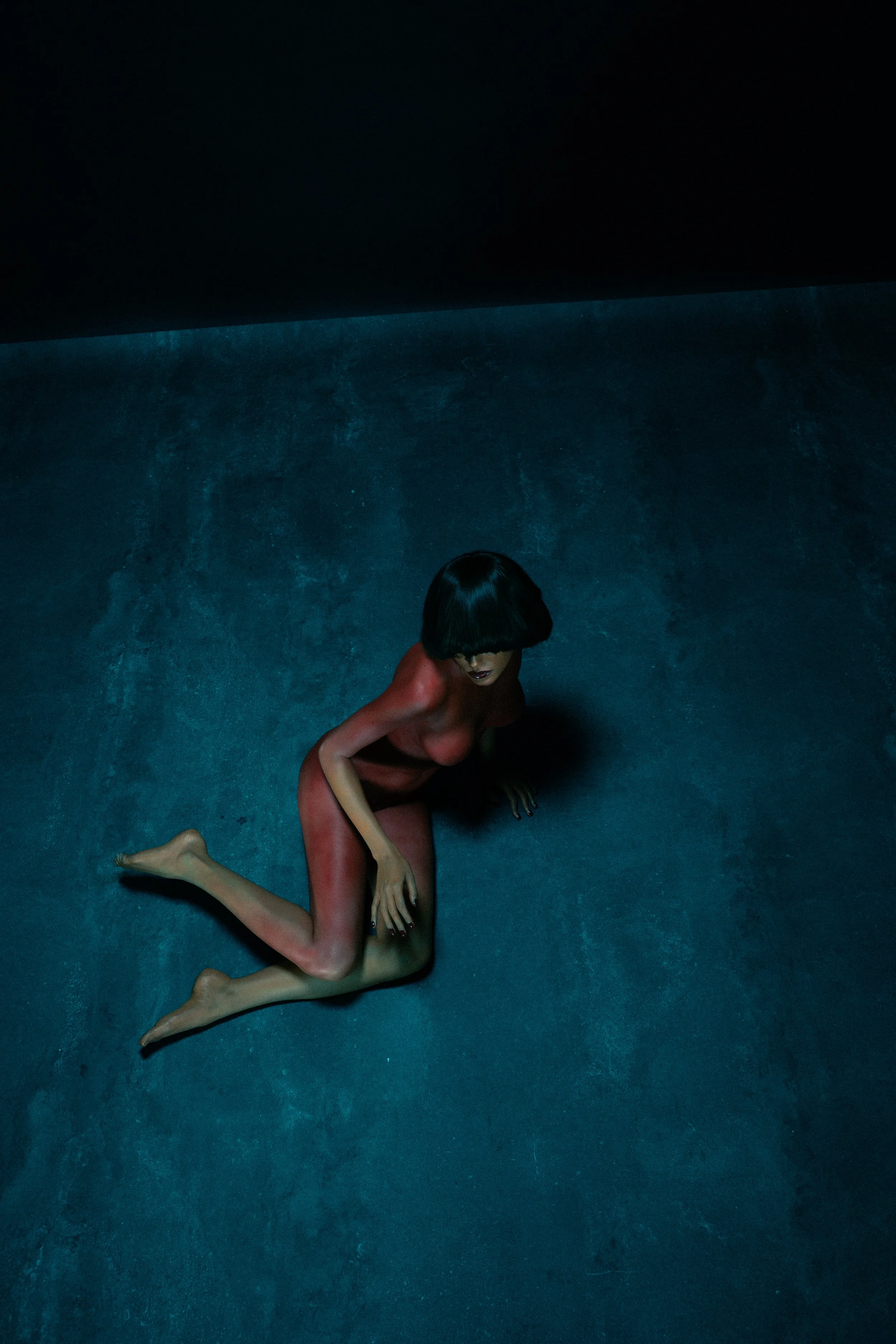 A person with dark hair and dark lipstick kneeling on a textured blue floor in a dark room, illuminated by a faint light from above.