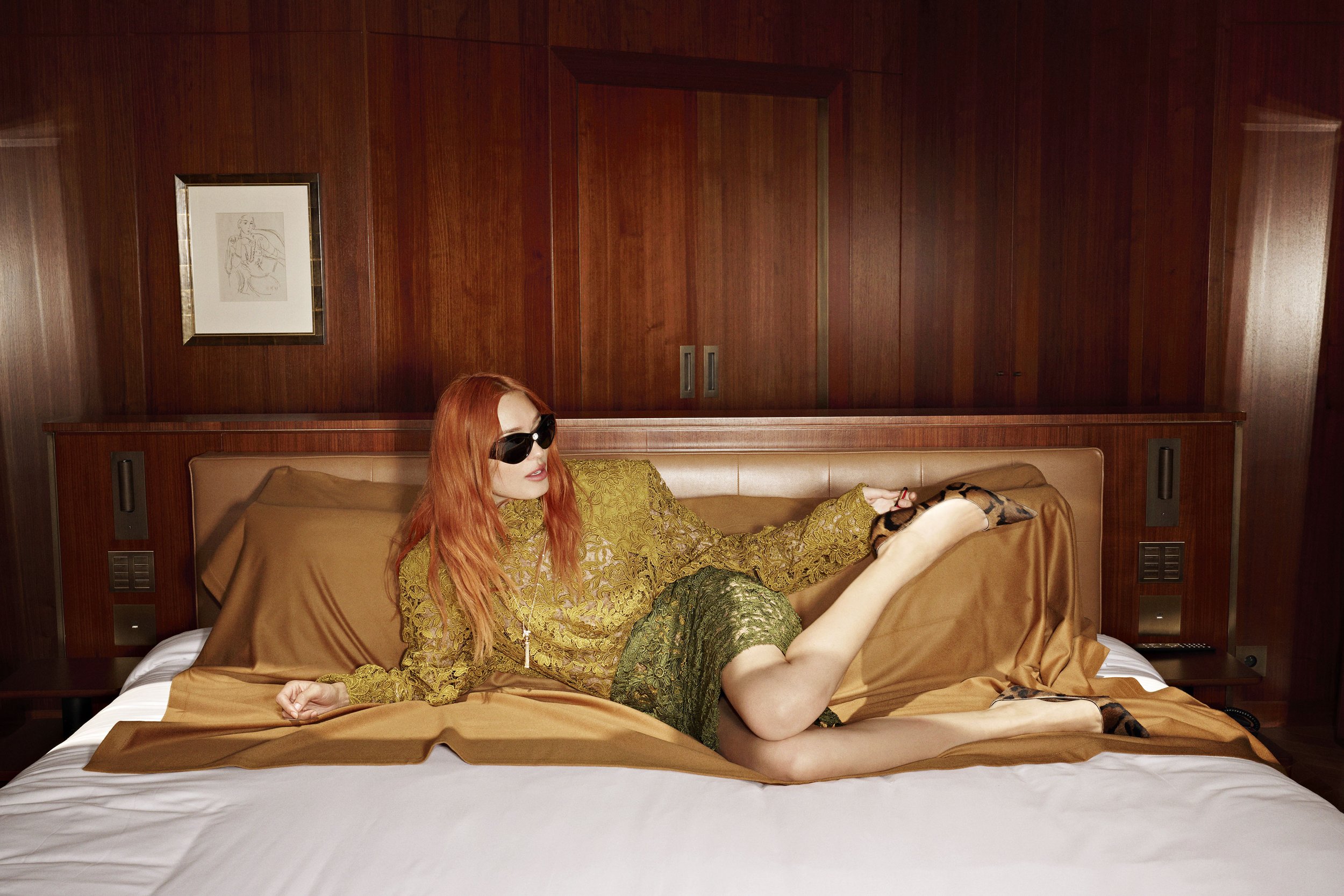 A woman with long red hair wearing sunglasses, a gold patterned top, and a green lace skirt lying on a bed in a room with wooden walls.