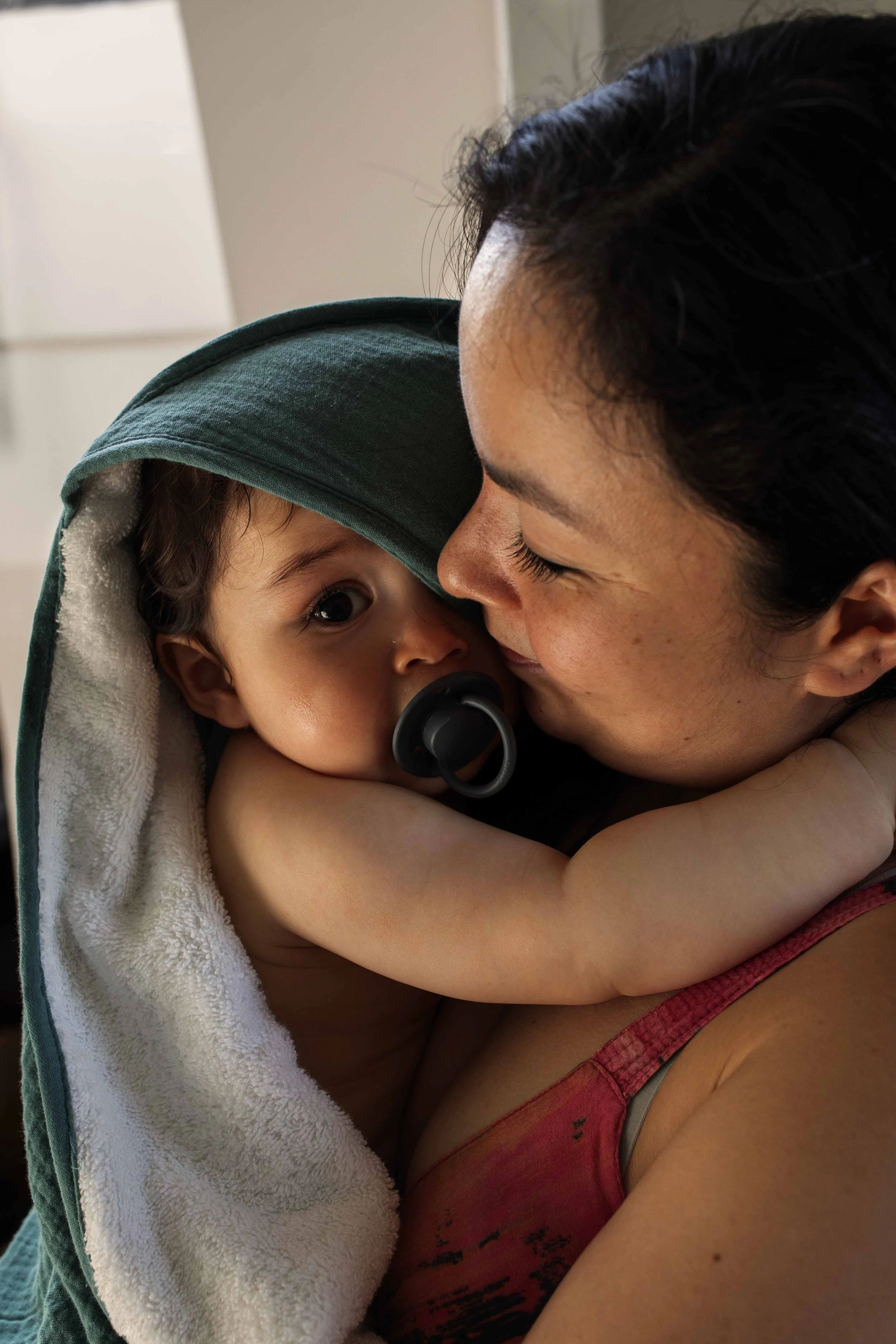 A woman holds a young child with a pacifier in his mouth, both embracing and touching noses, in a close-up indoor photo.