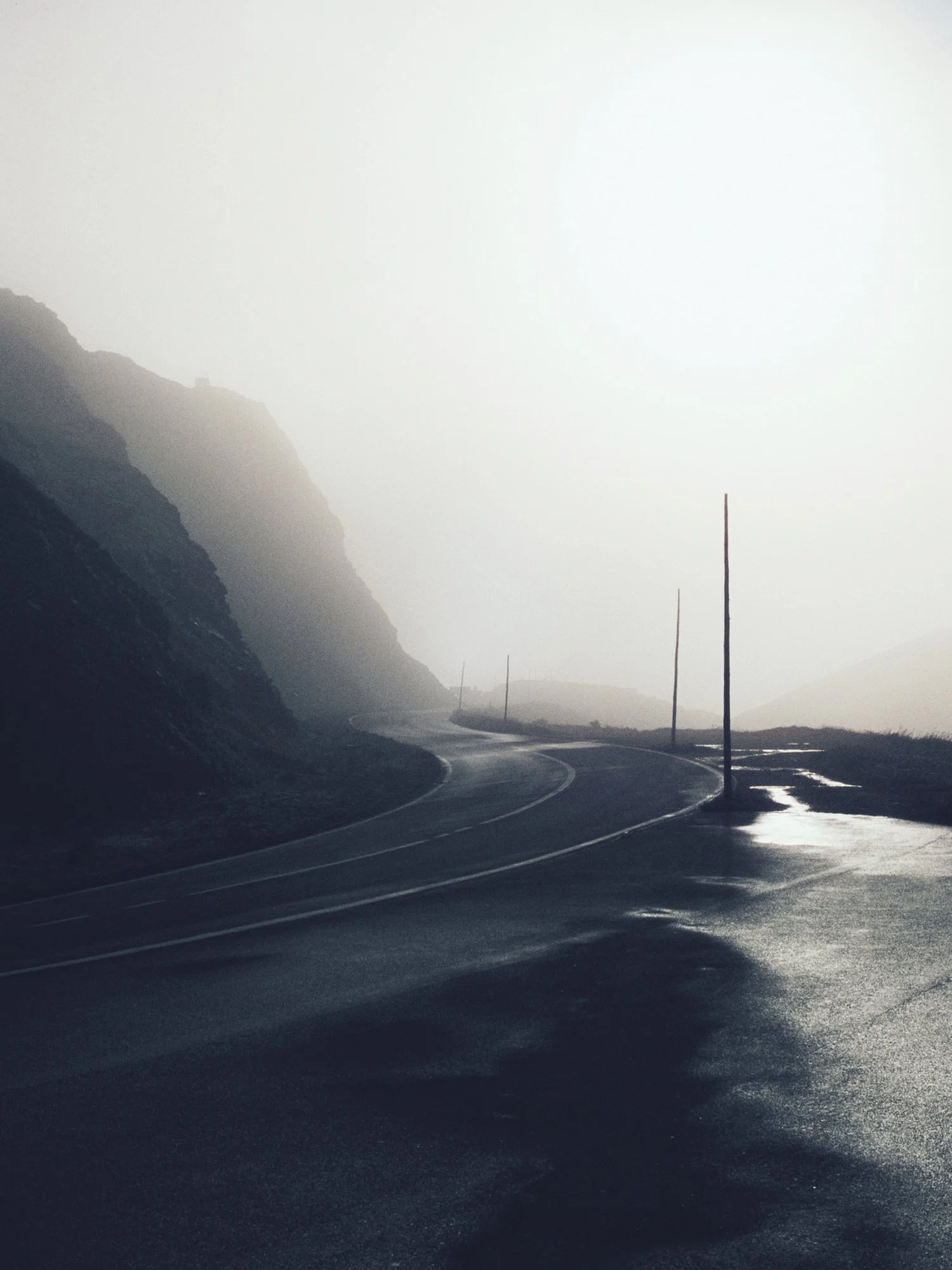 A winding road on a foggy, mountainous landscape with wet pavement and partially visible utility poles along the roadside.