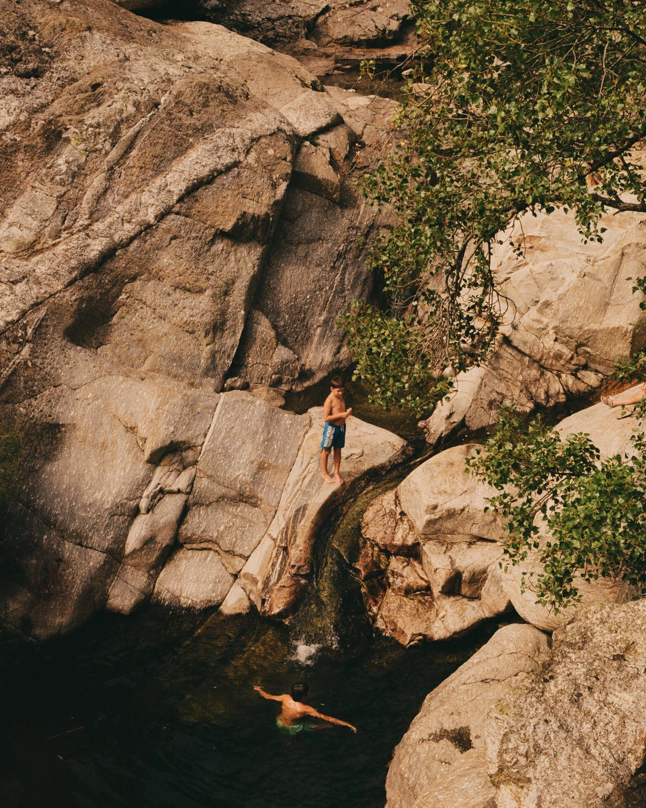 A boy in blue swim trunks standing on a rock ledge above a small stream, with another boy swimming in the water below among large rocks and surrounded by trees.