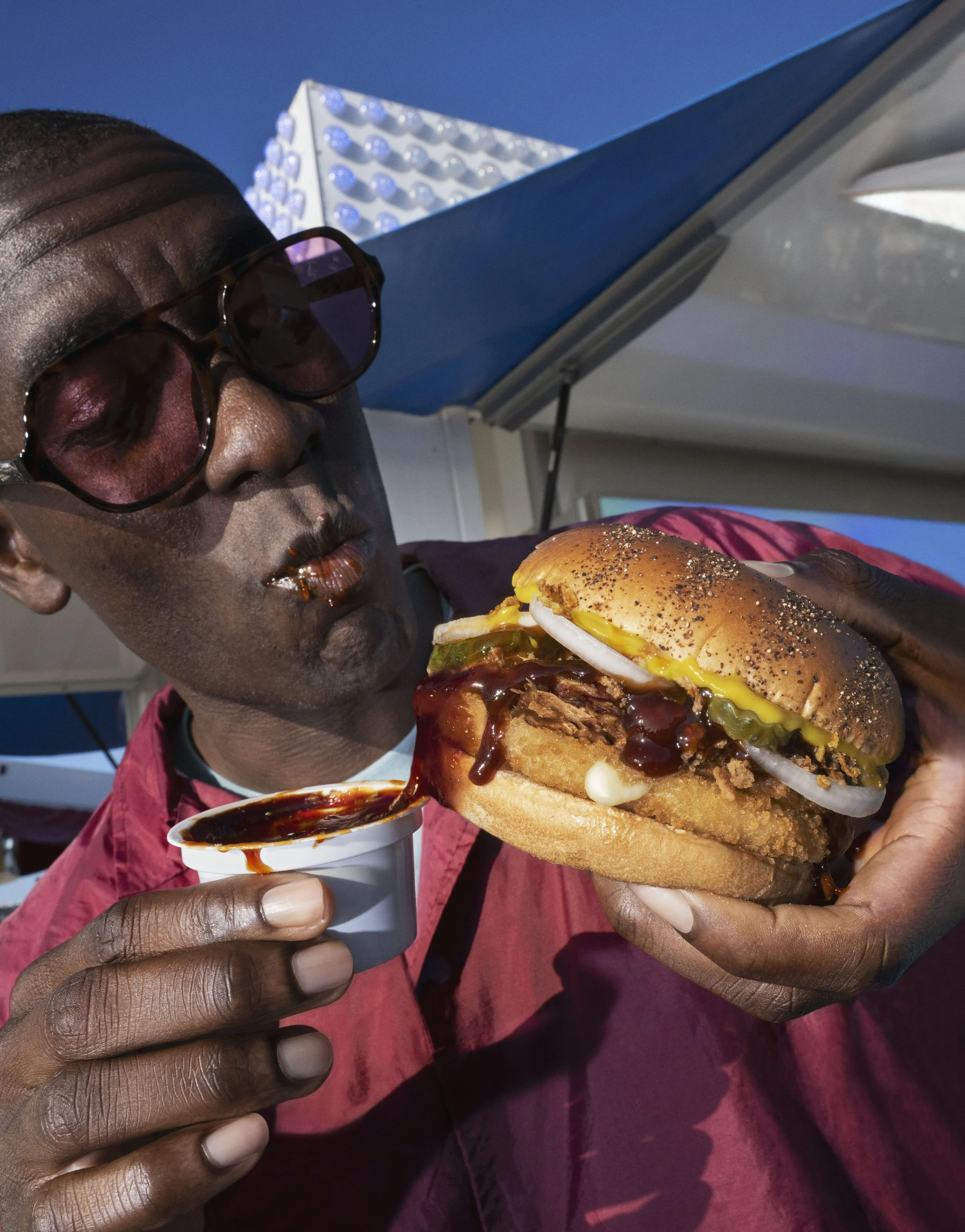 A man wearing sunglasses holding a large cheeseburger with fried chicken, pickles, onion, and sauce. He is about to dip fries into a small container of ketchup.