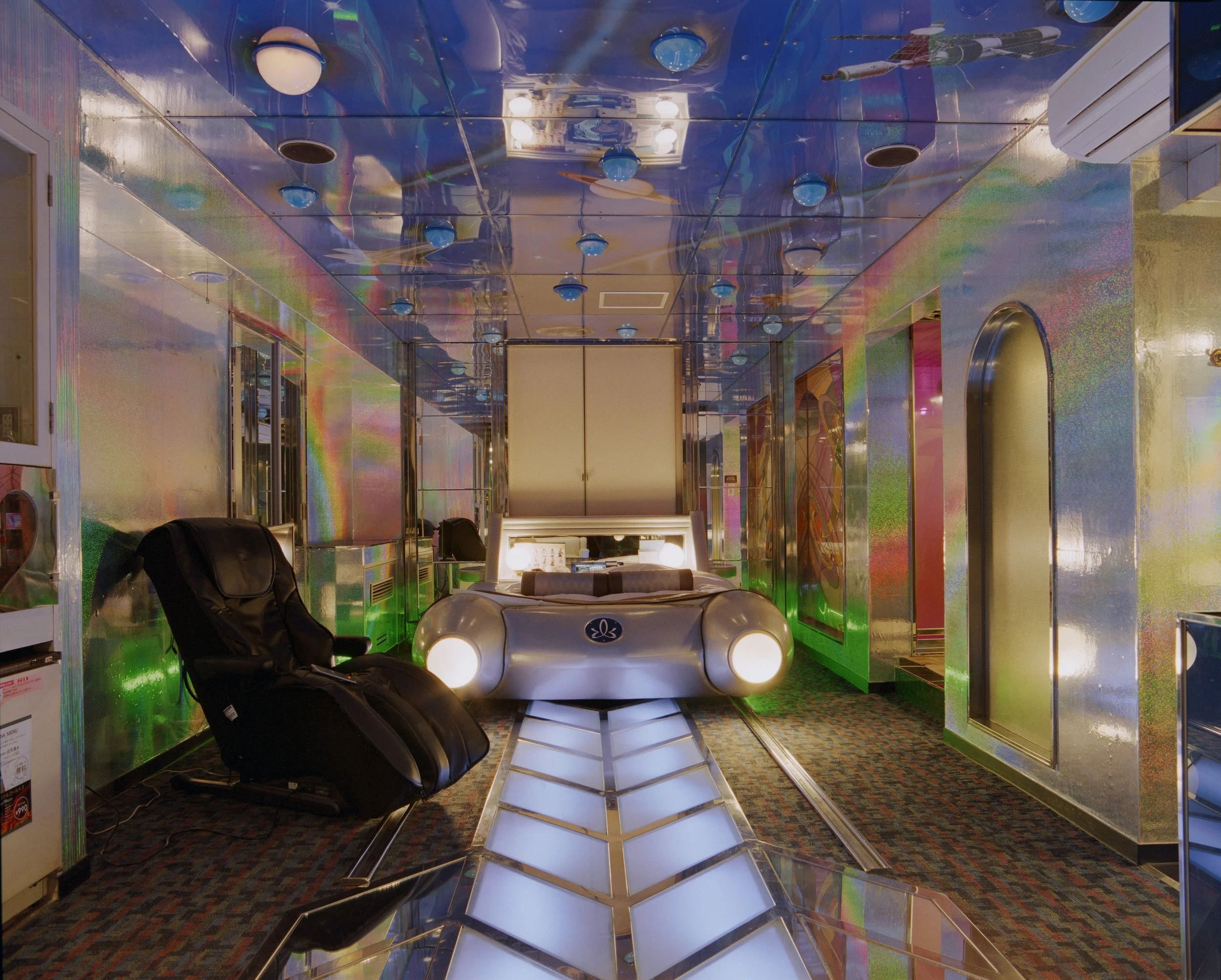 A room with metallic iridescent walls and ceiling, featuring blue ceiling fans and multiple round white lights. There is a black massage chair on the left, a futuristic silver bed with head and tail lights in the center, and colorful abstract artwork