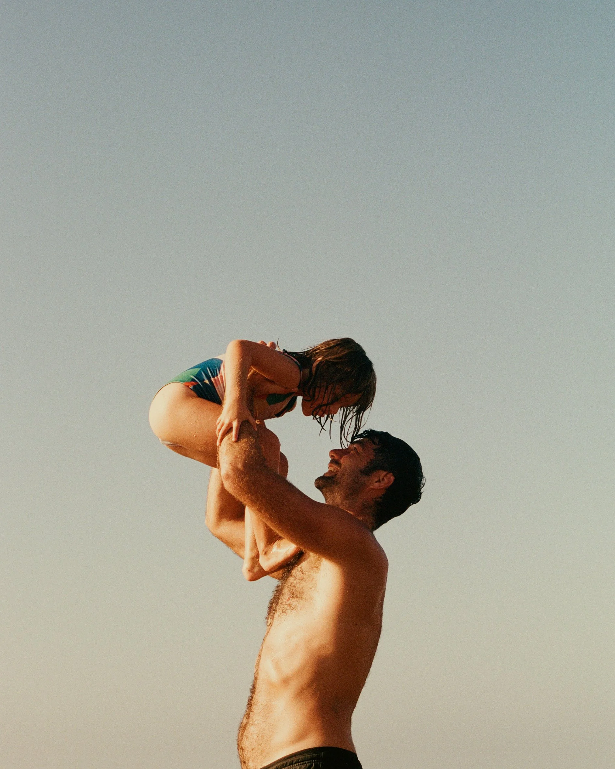 A shirtless man lifting a woman in a swimsuit in the air against a clear sky