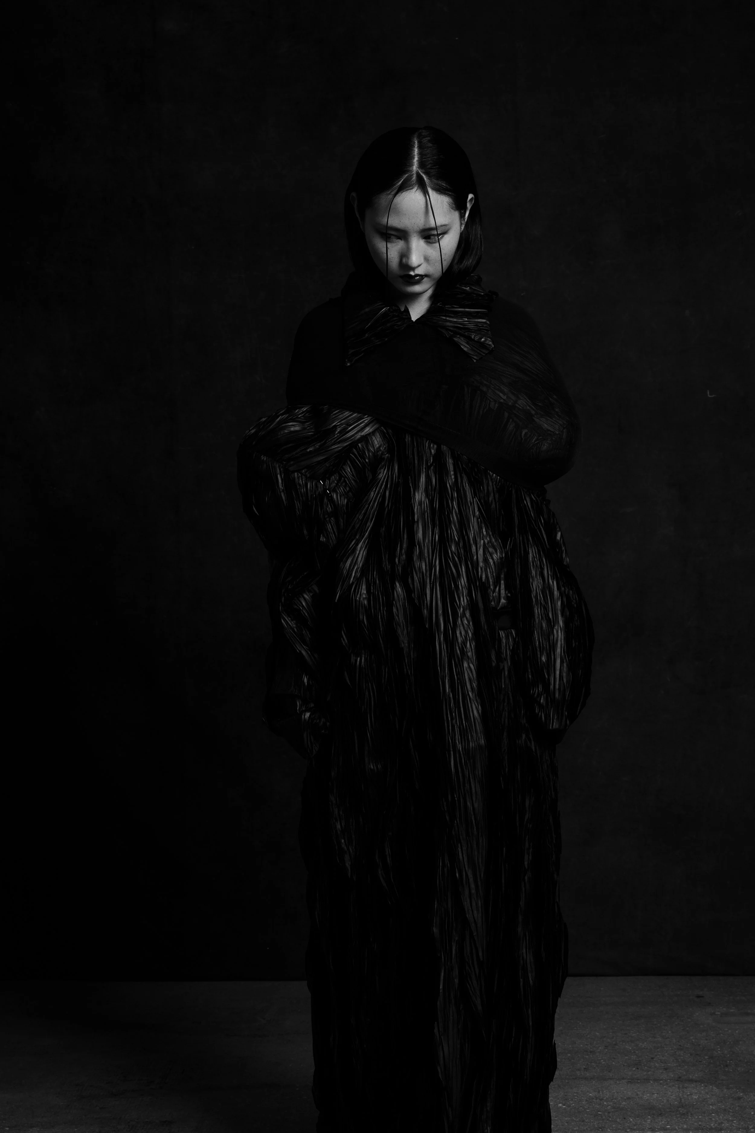 A woman with short dark hair stands against a dark background, wearing a black, shiny, textured outfit with a large collar, looking downward with a somber expression.