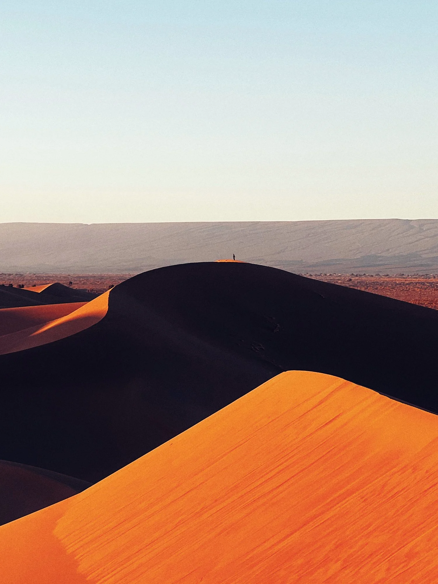 Sand dunes in a desert during sunset, with a person standing on the highest dune in the distance.