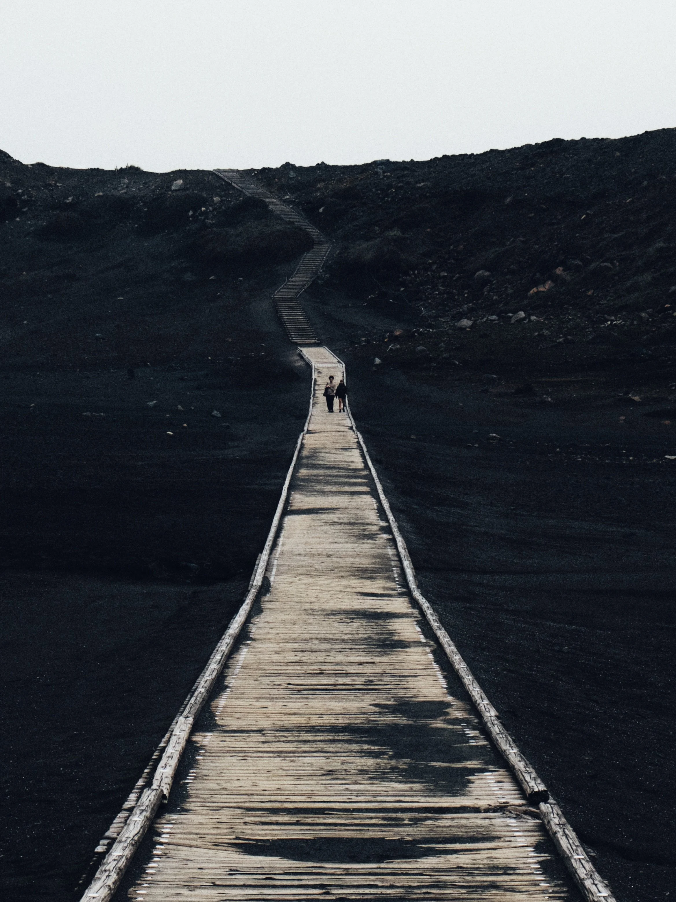 A long wooden footbridge extending over dark, barren volcanic landscape, with two people walking in the distance under an overcast sky.