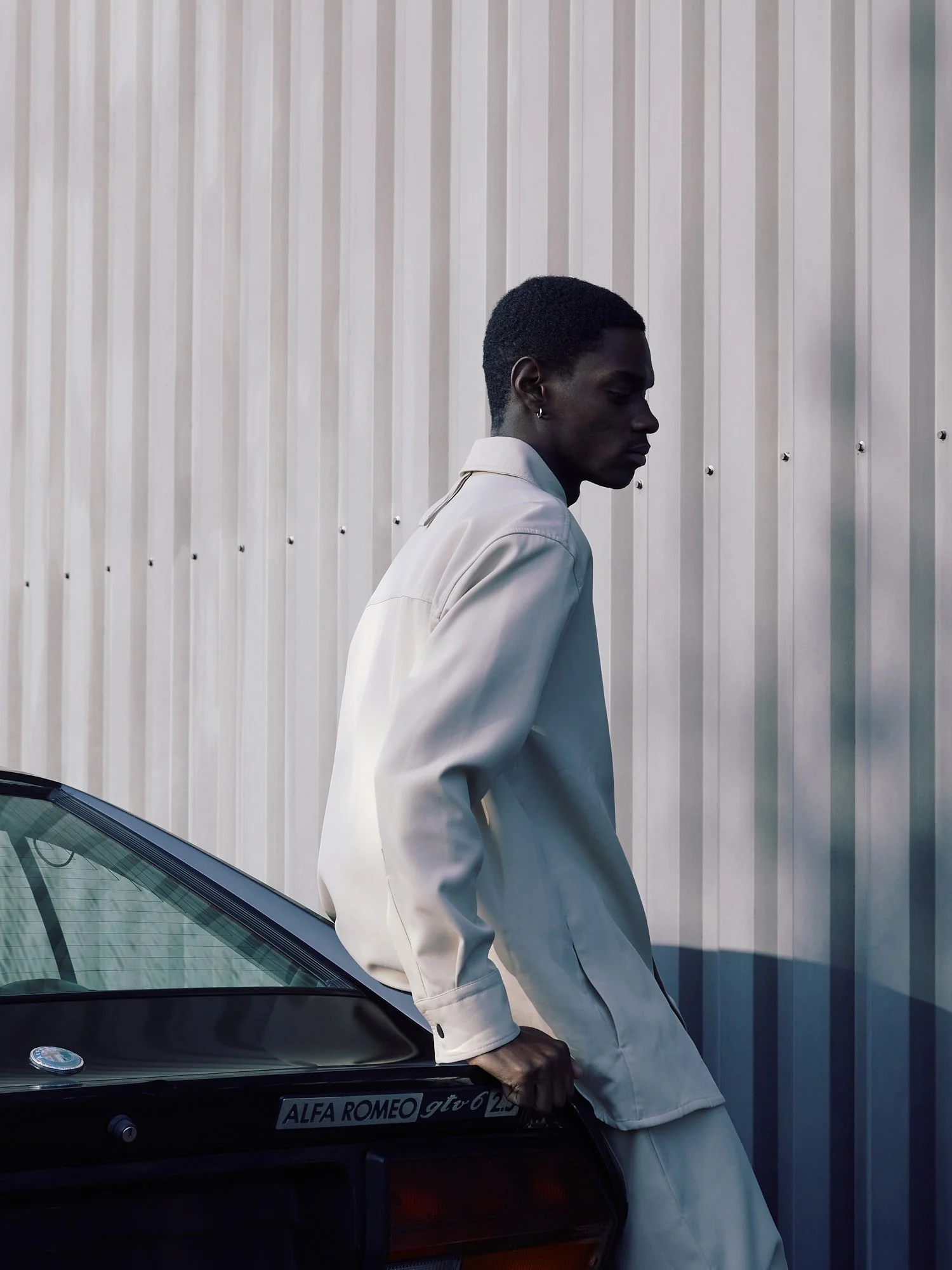 A young man with dark skin leaning against a black vintage Alfa Romeo GTV 2.5 car, with hands in pockets, standing in front of a light-colored corrugated metal wall.
