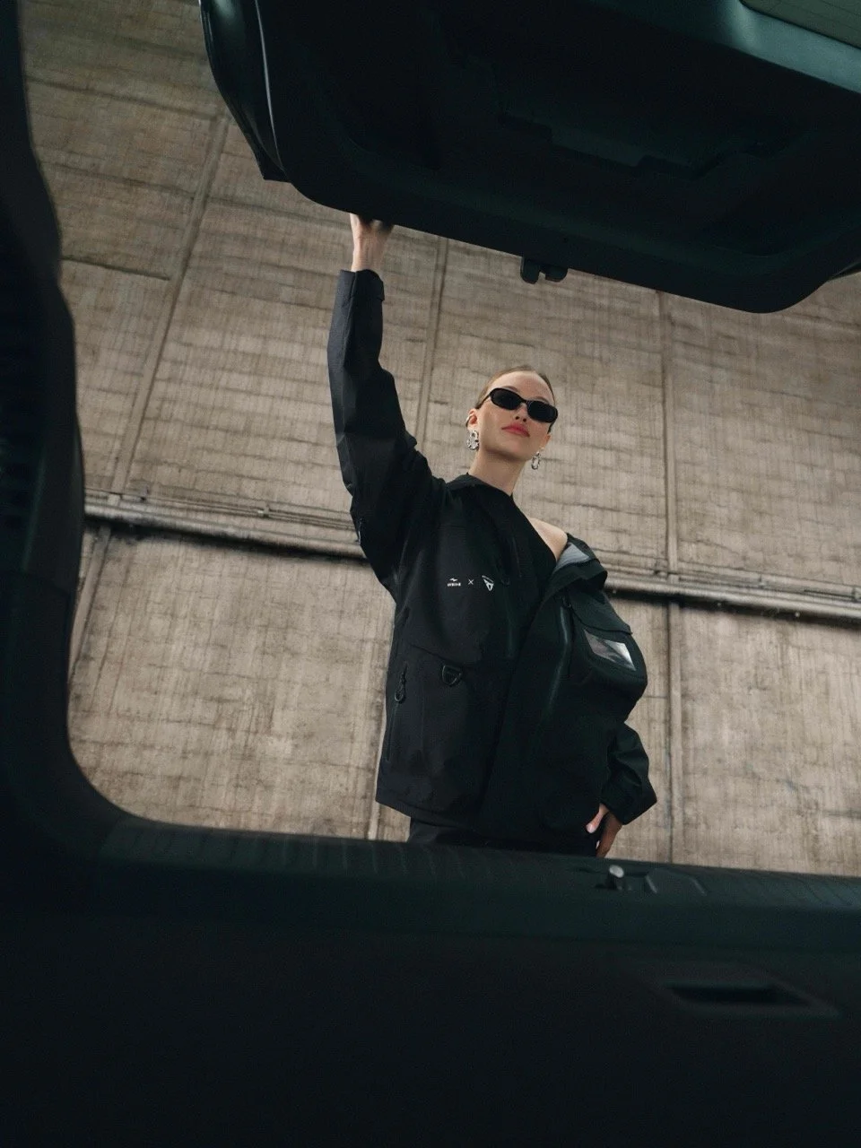 A woman with short hair wearing sunglasses, earrings, a black jacket, and a black bag, standing in a parking garage, viewed from the trunk of a car.