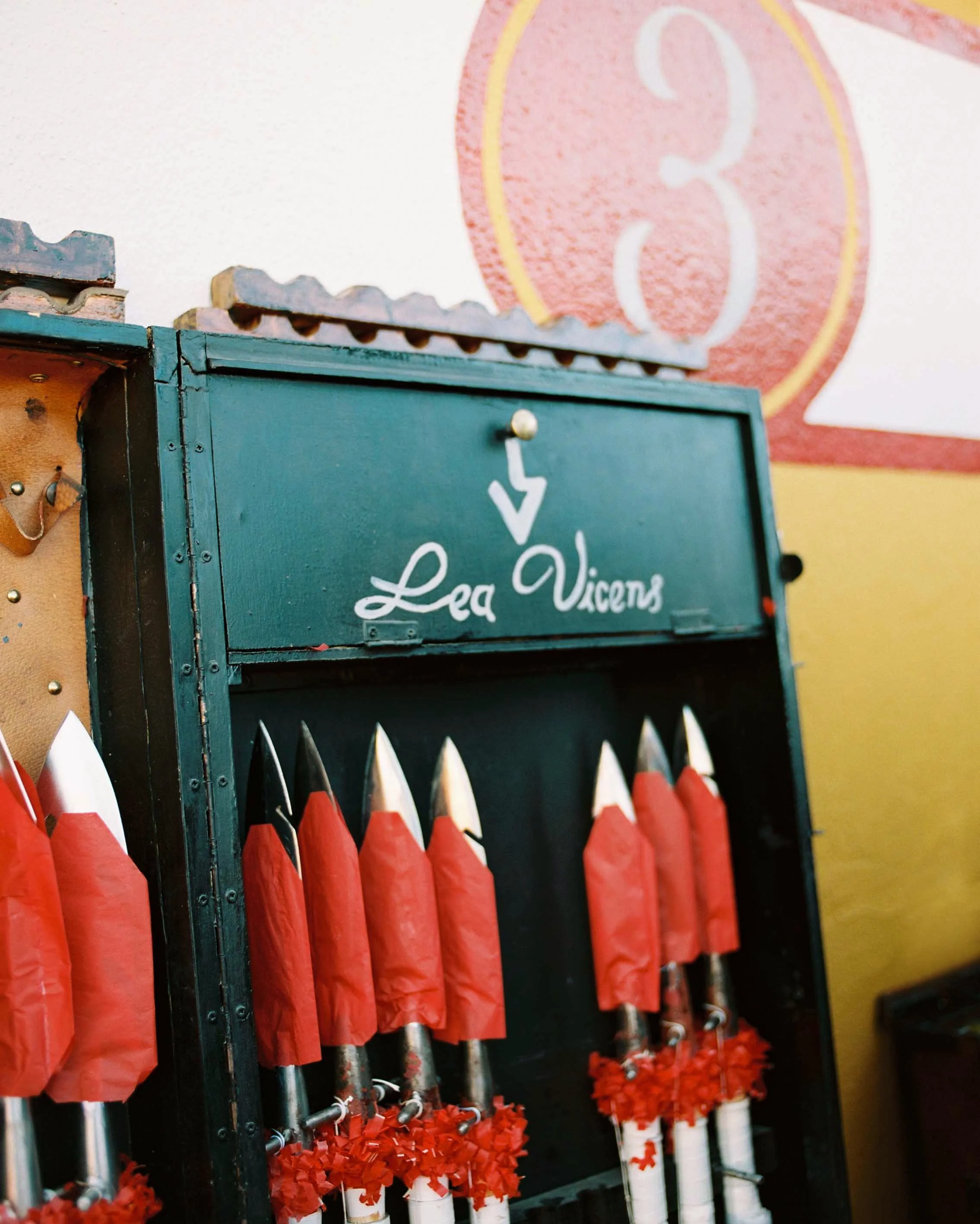 A display of red and white fireworks rockets inside a dark green box with the words "Lea Vicens" written on it, set against a colorful wall.